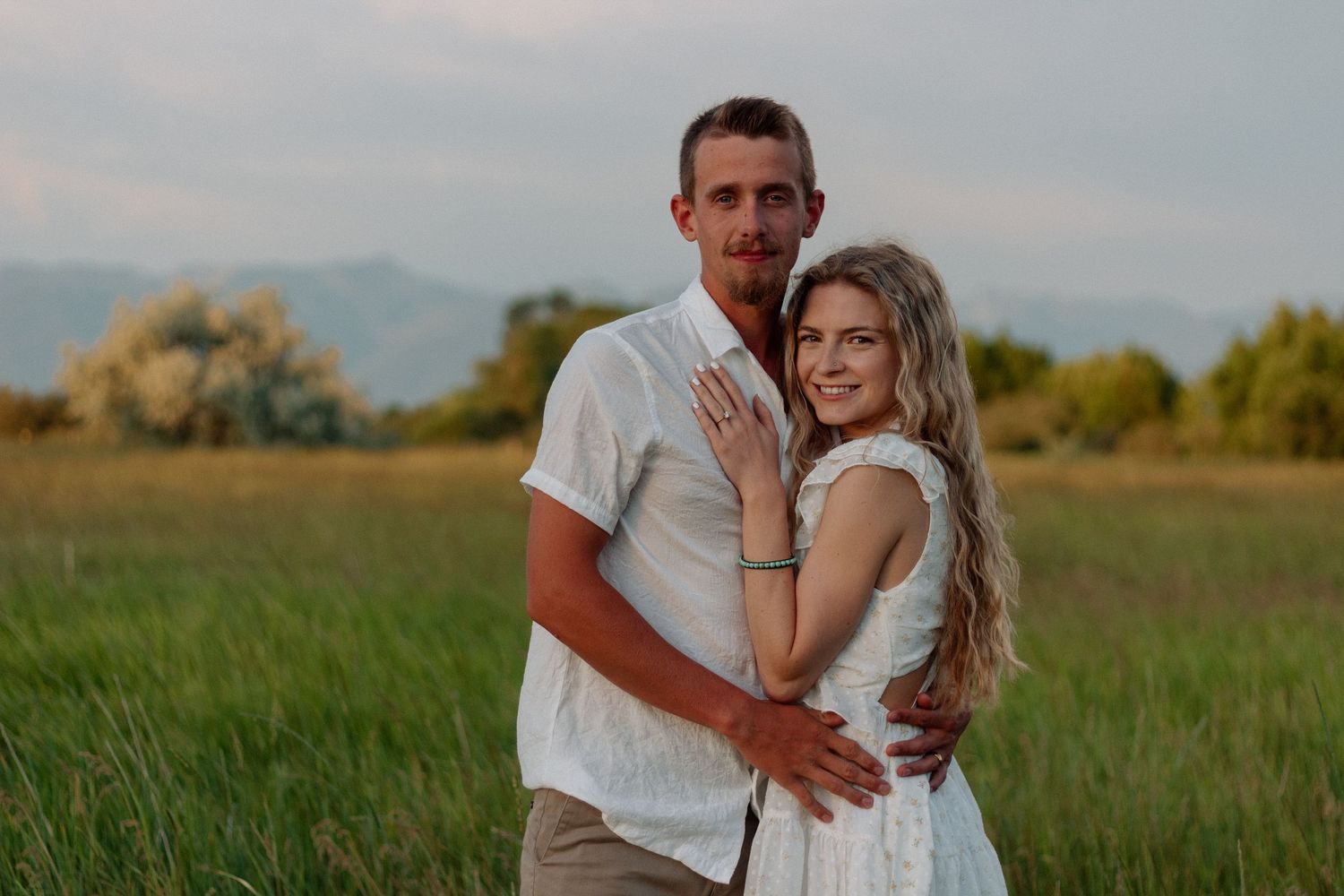 Couple embracing in a rustic field with mountains in the background during golden hour.