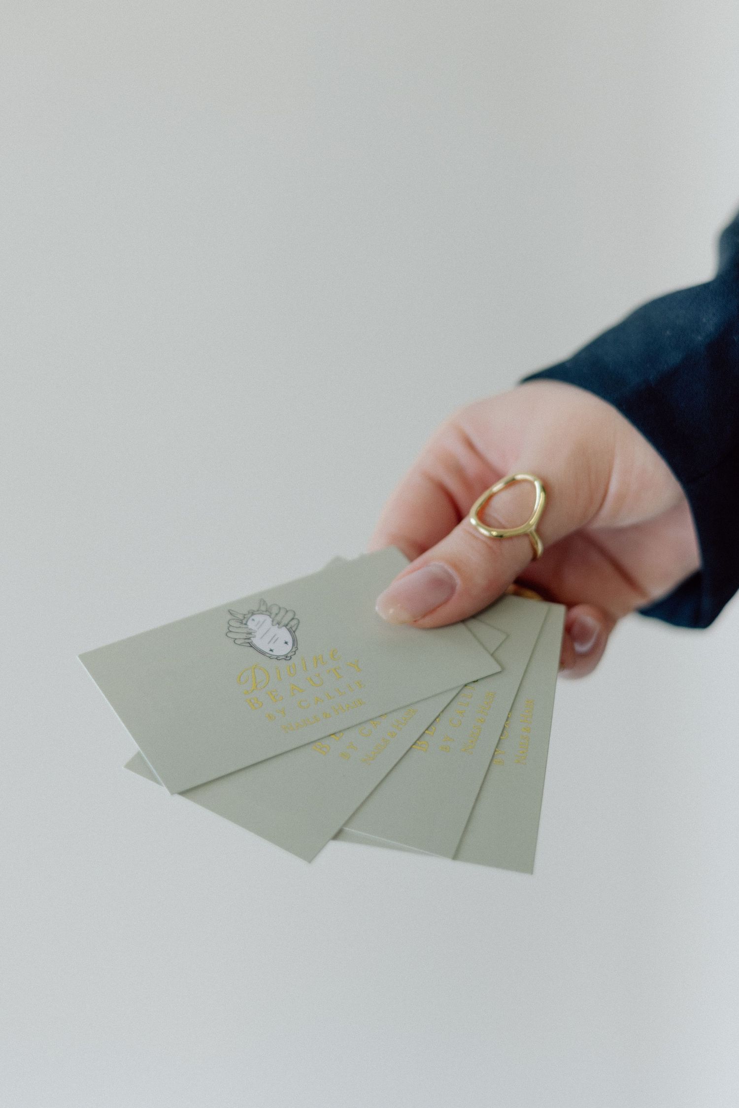 A hand wearing a gold hexagonal ring holds minimalist gray business cards with a logo design.