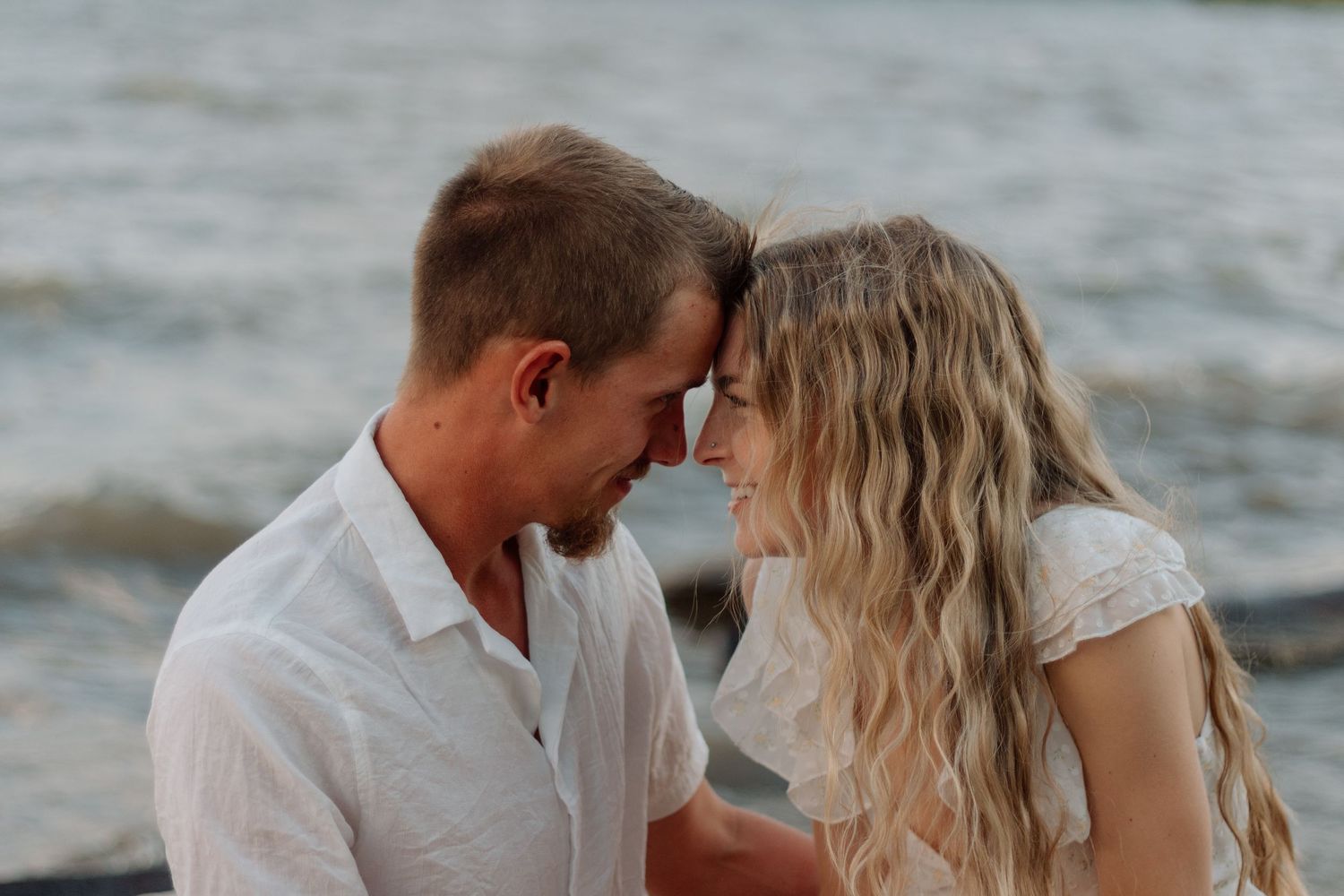 Romantic couple in white outfits share tender moments during a beach photoshoot at sunset.