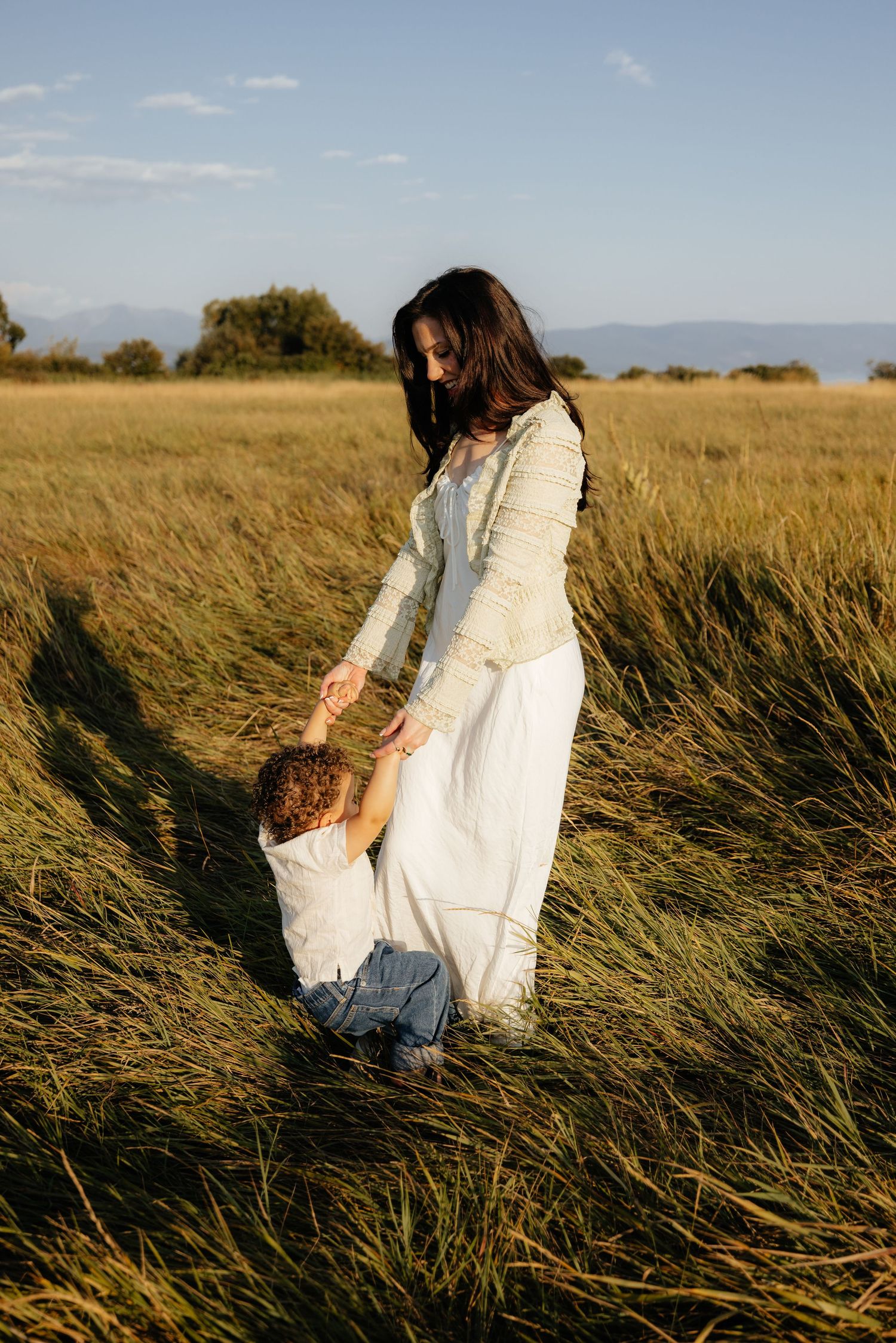 Two people in white dresses embrace while standing in a golden wheat field at sunset.