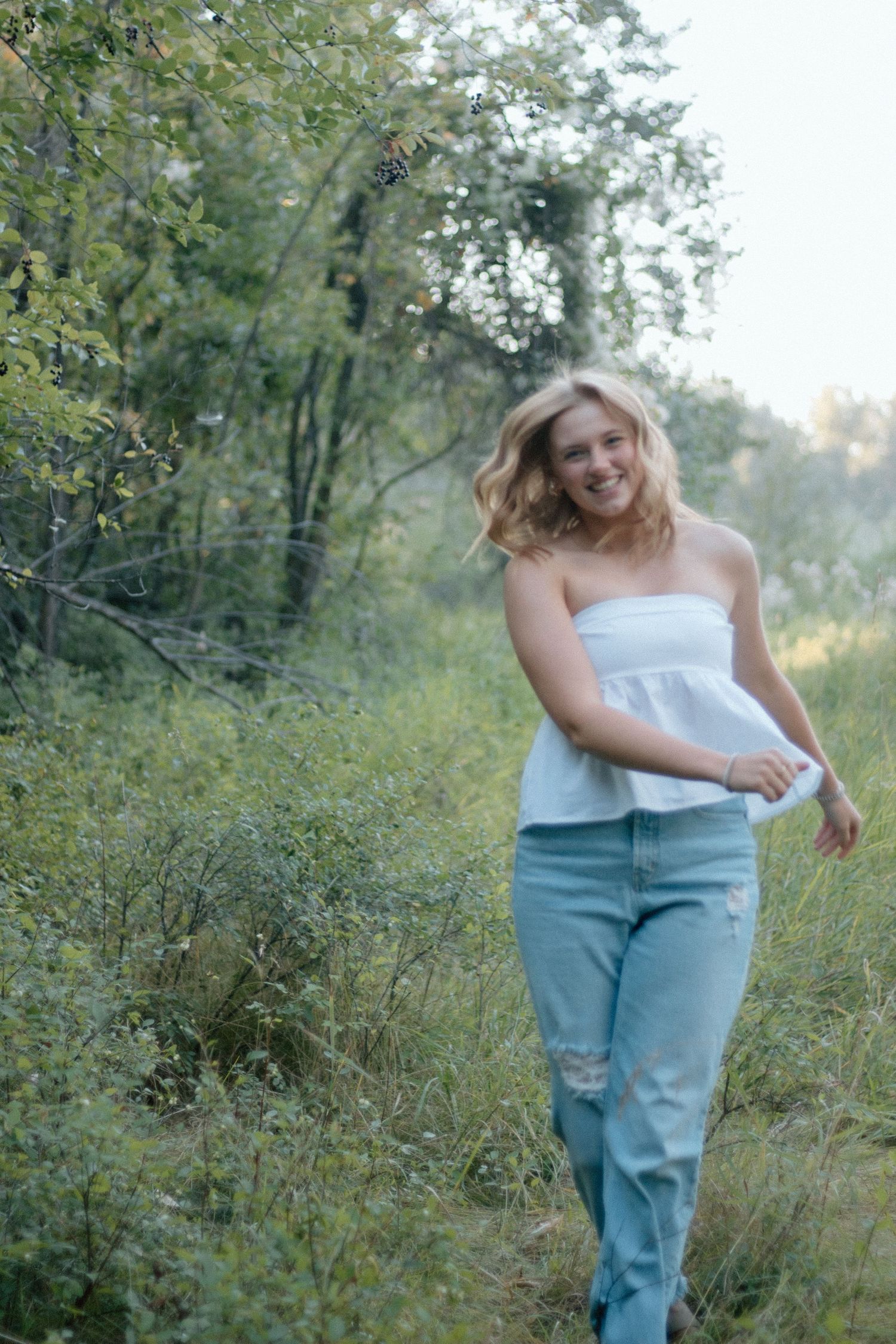 Person in light top and blue jeans poses playfully in a natural outdoor setting with green foliage.