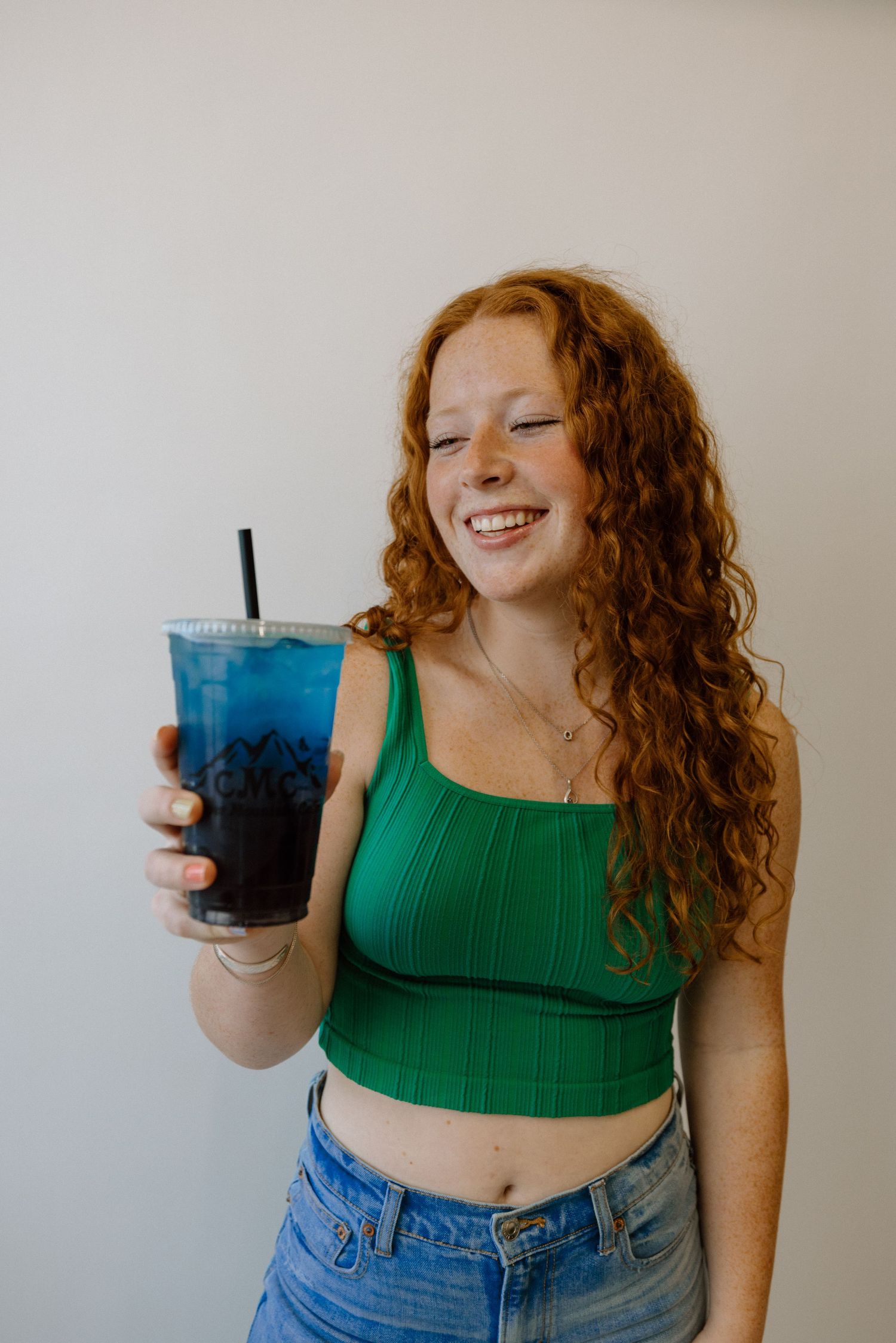 Person in green crop top and jeans holds blue drink while posing against white background.