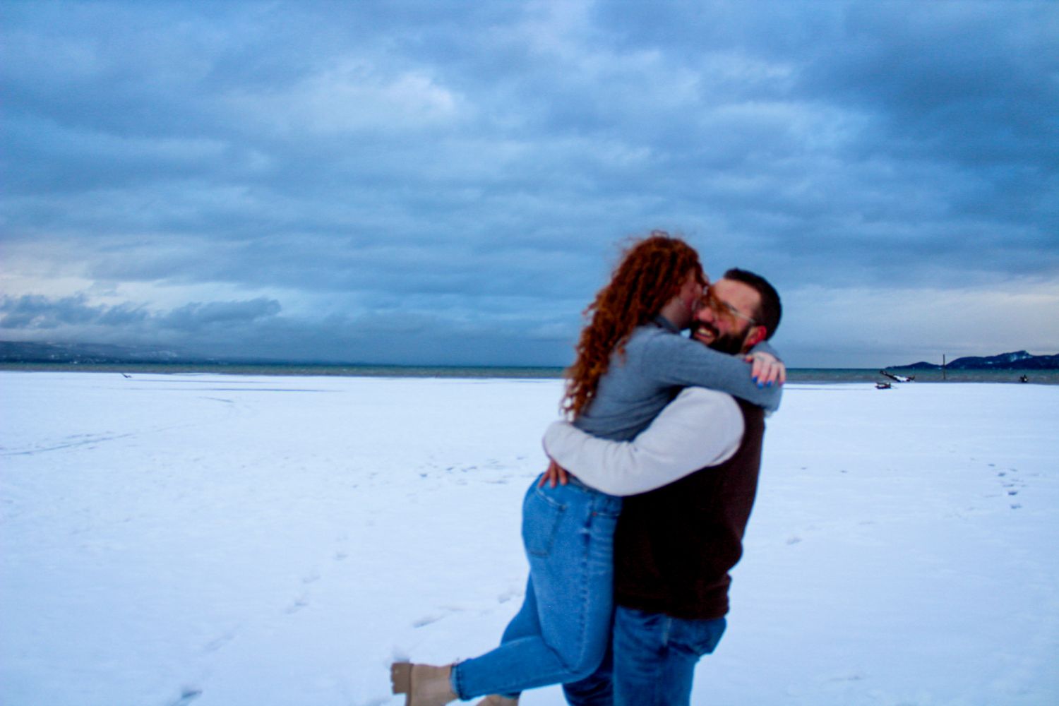 A couple embraces on a snow-covered landscape with dramatic cloudy skies in the background.