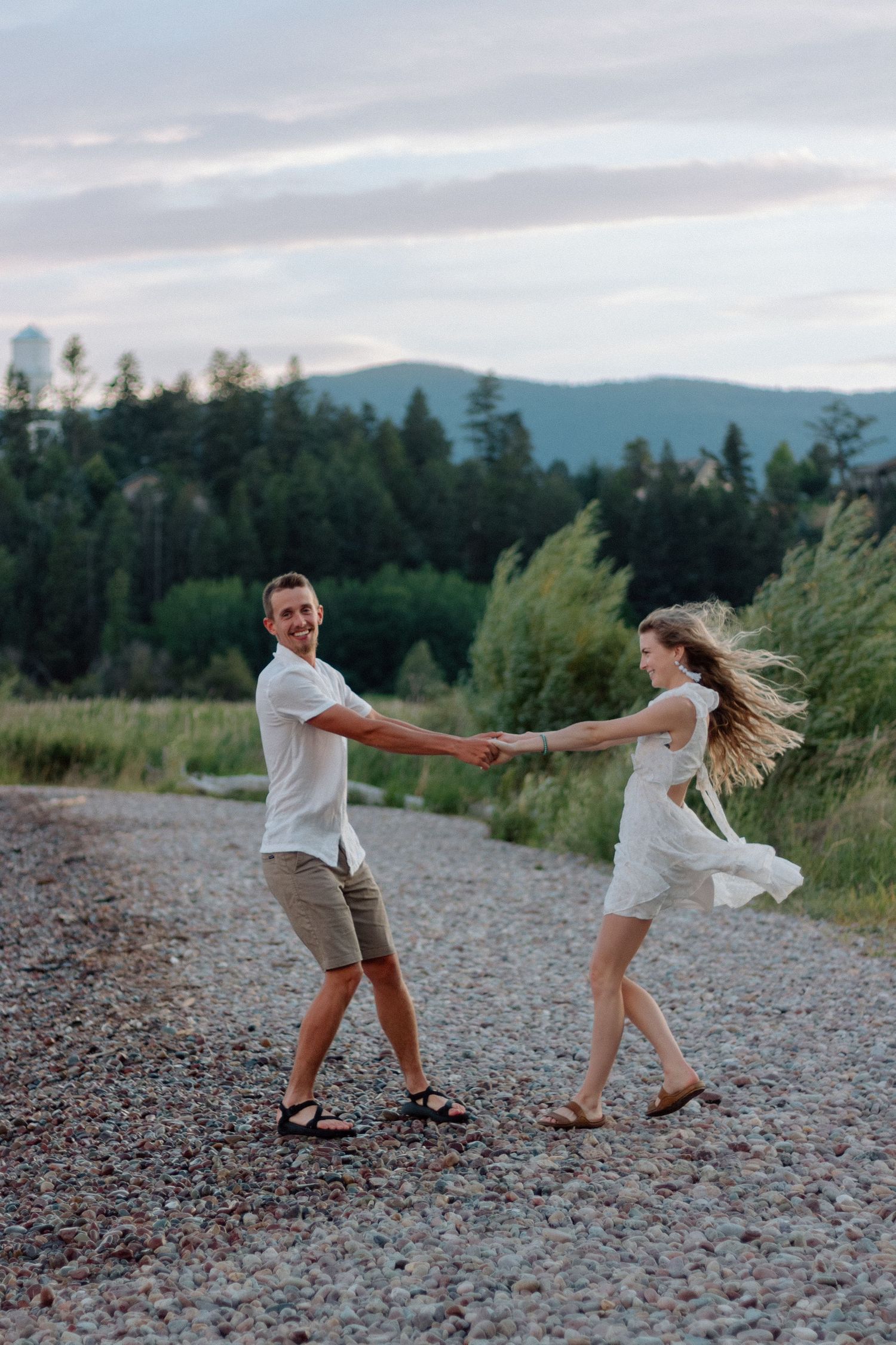 A playful couple spins and dances together on a gravel path with mountains and forest in the background.