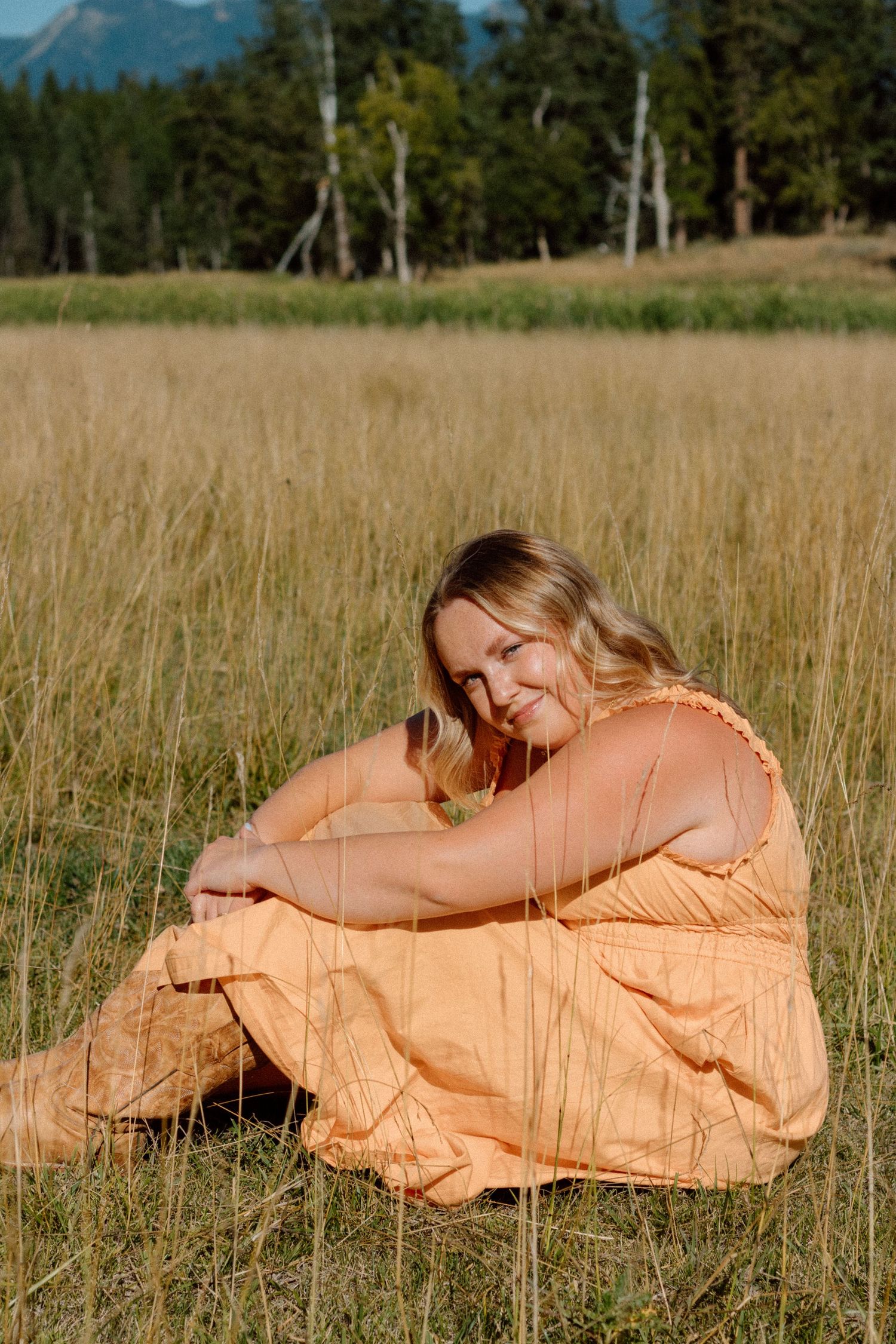 A person in a flowing peach-colored dress lies peacefully in a golden grass field with mountains in the background.