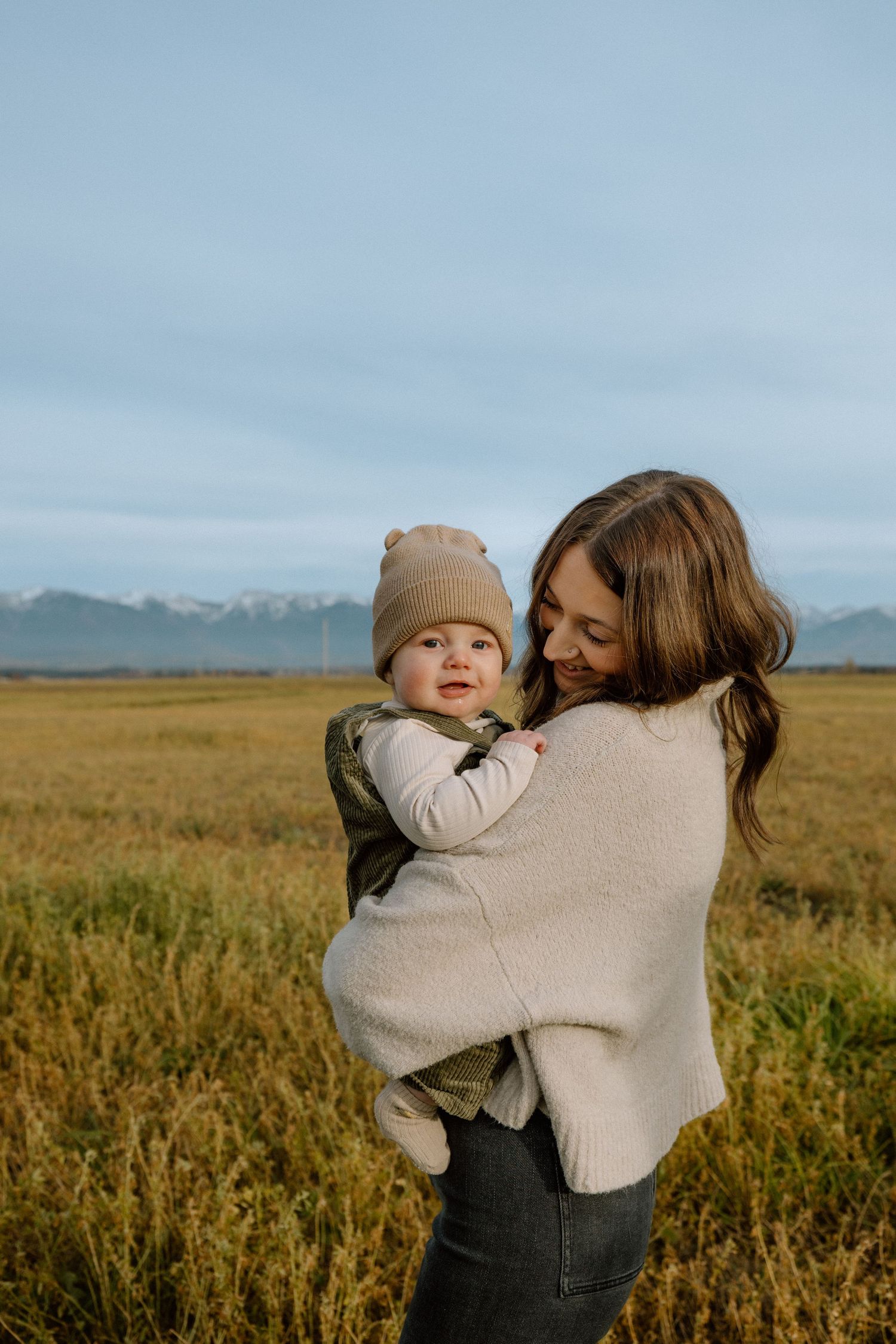 A tender family moment captured in a golden field with snow-capped mountains visible in the distance.
