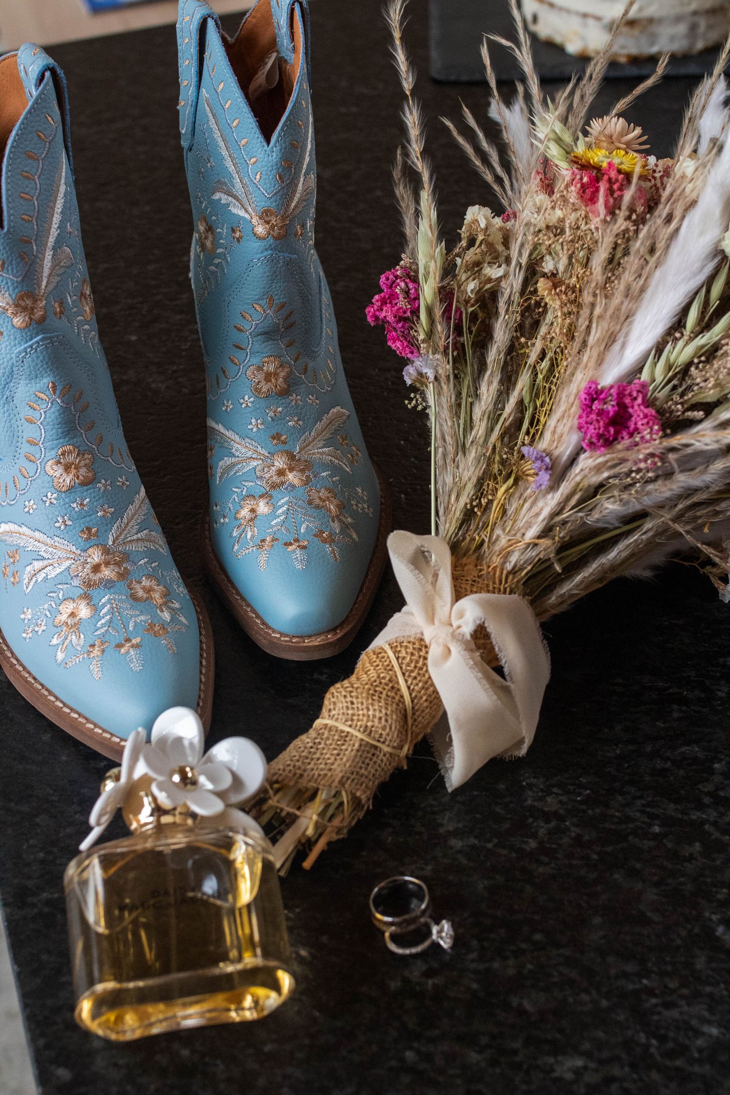 Light blue cowboy boots with floral embroidery displayed with dried flowers and glass bottles on dark background.