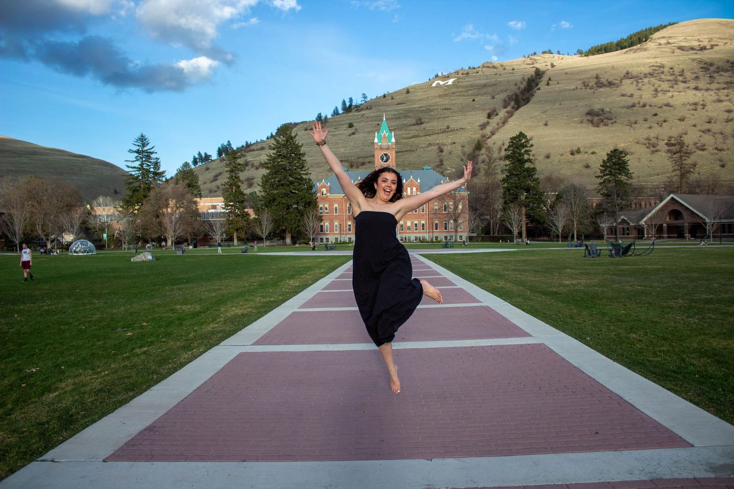 Graduate jumping for joy on campus walkway with mountains in background.