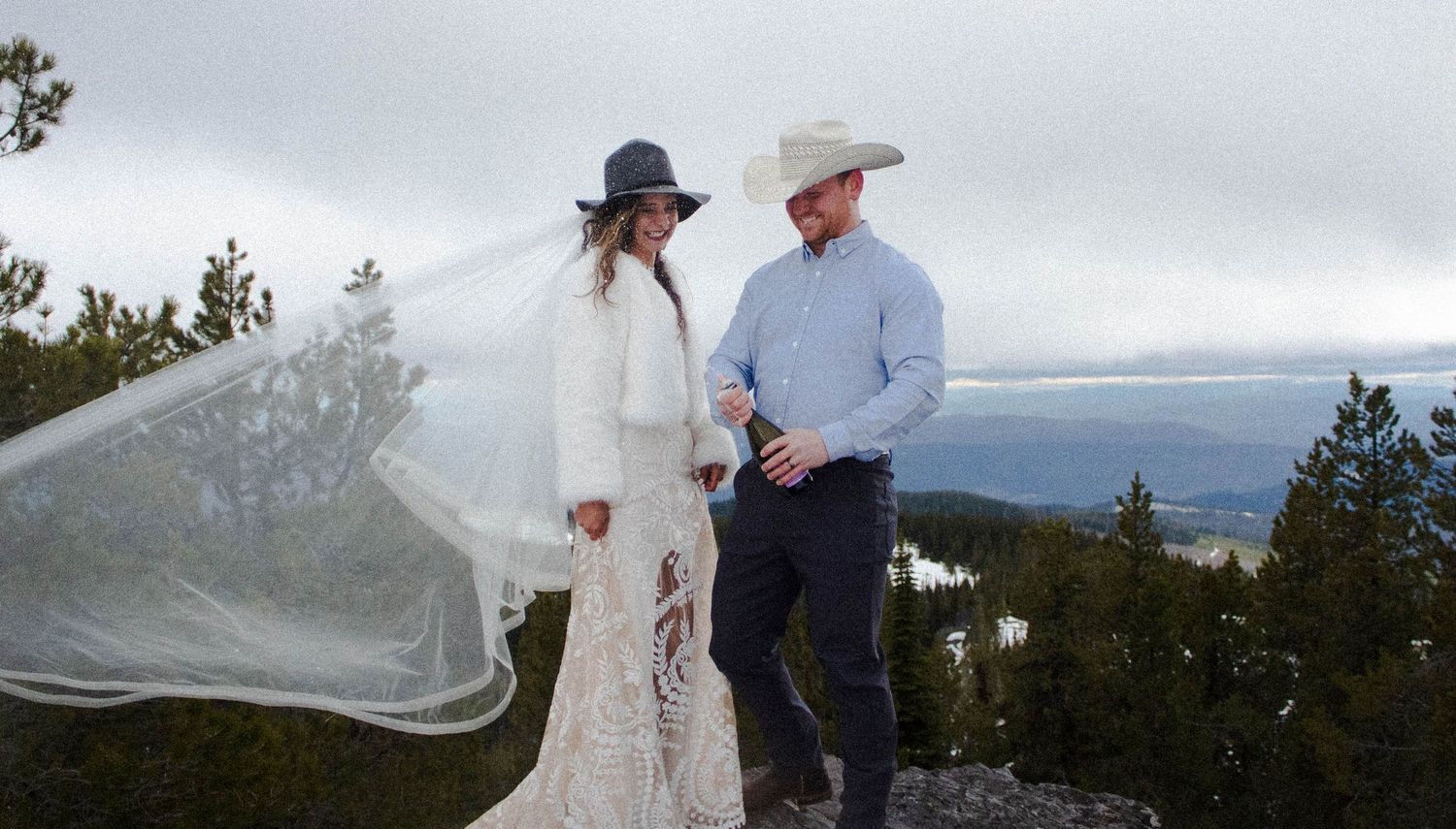 A couple in wedding attire poses together on a scenic mountain overlook with a flowing veil in the breeze.