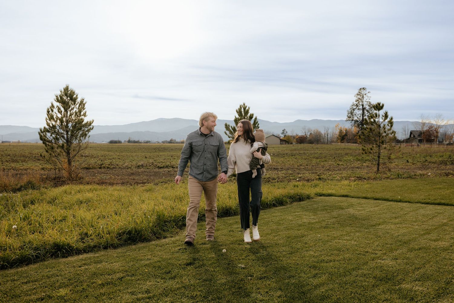A couple walks together through a scenic grassland landscape with mountains visible in the background on a cloudy day.