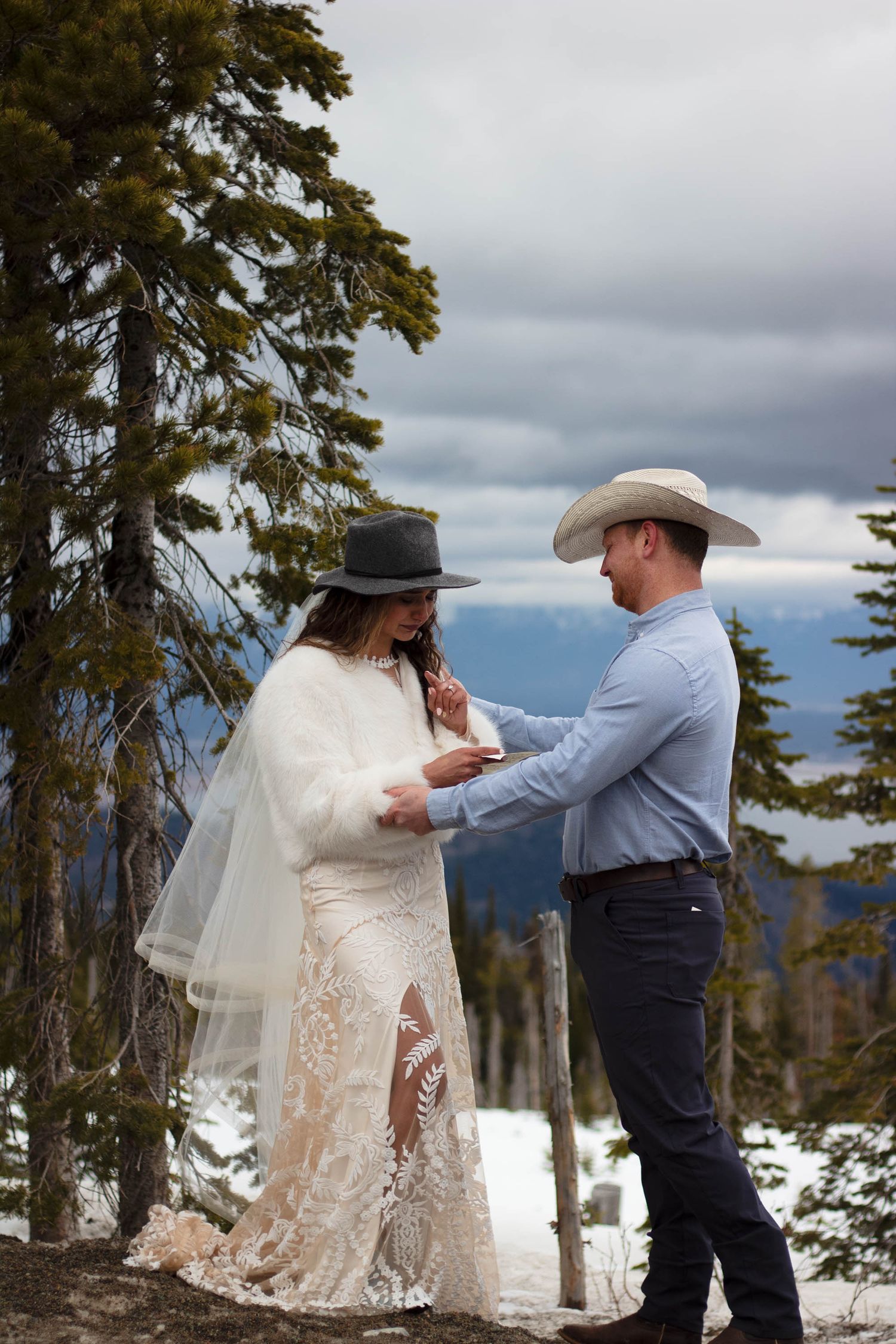 A couple dances together in the snow-covered mountains with evergreen trees and cloudy skies in the background.