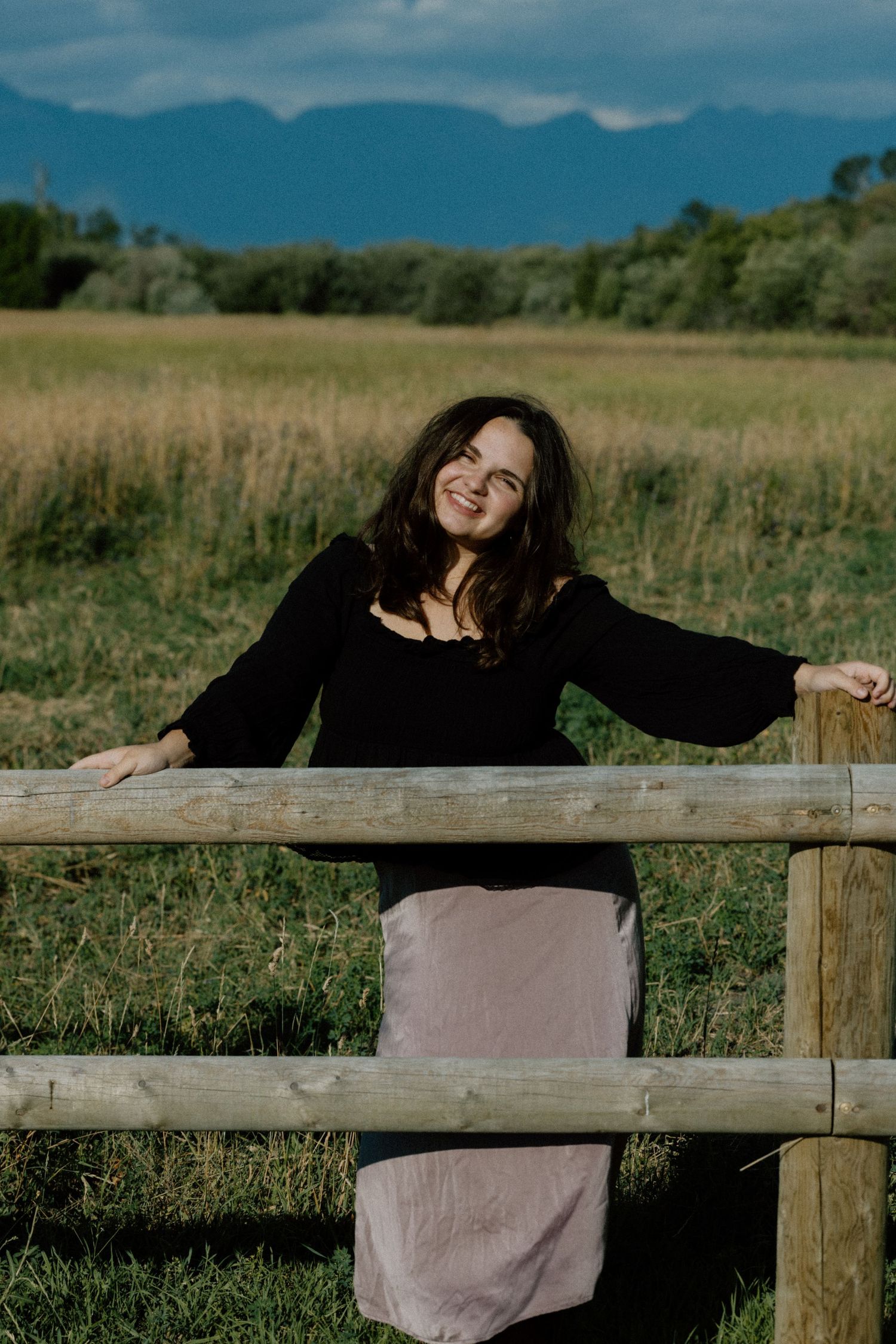 Figure in black leaning on wooden fence with mountain landscape in background.