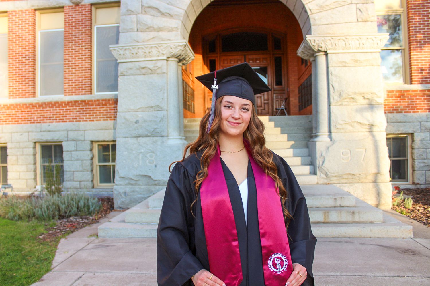 Graduate in cap and gown stands in front of collegiate stone building entrance during commencement ceremony.