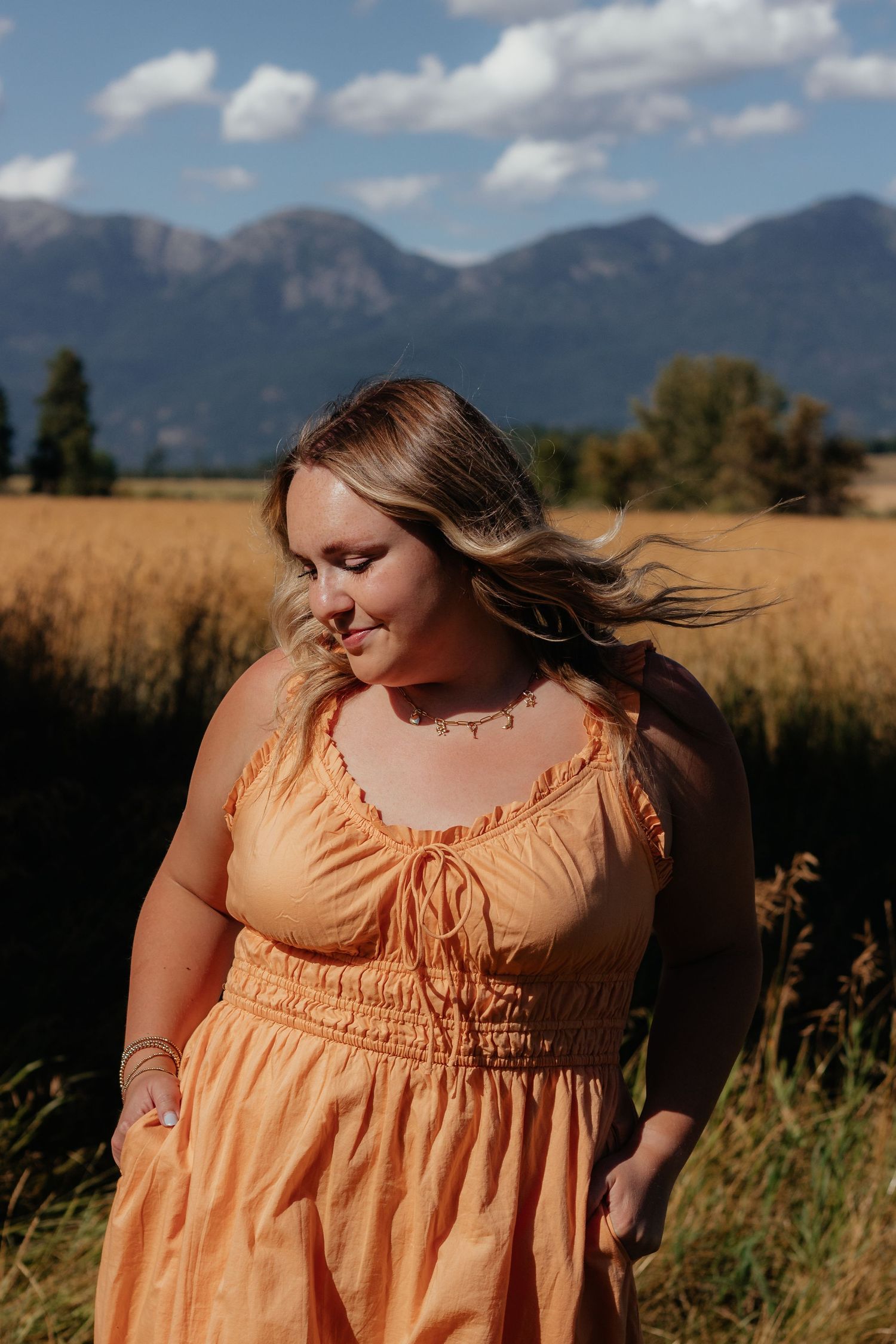 Soft peach-colored dress twirls in sunlit field with dramatic mountain landscape behind.