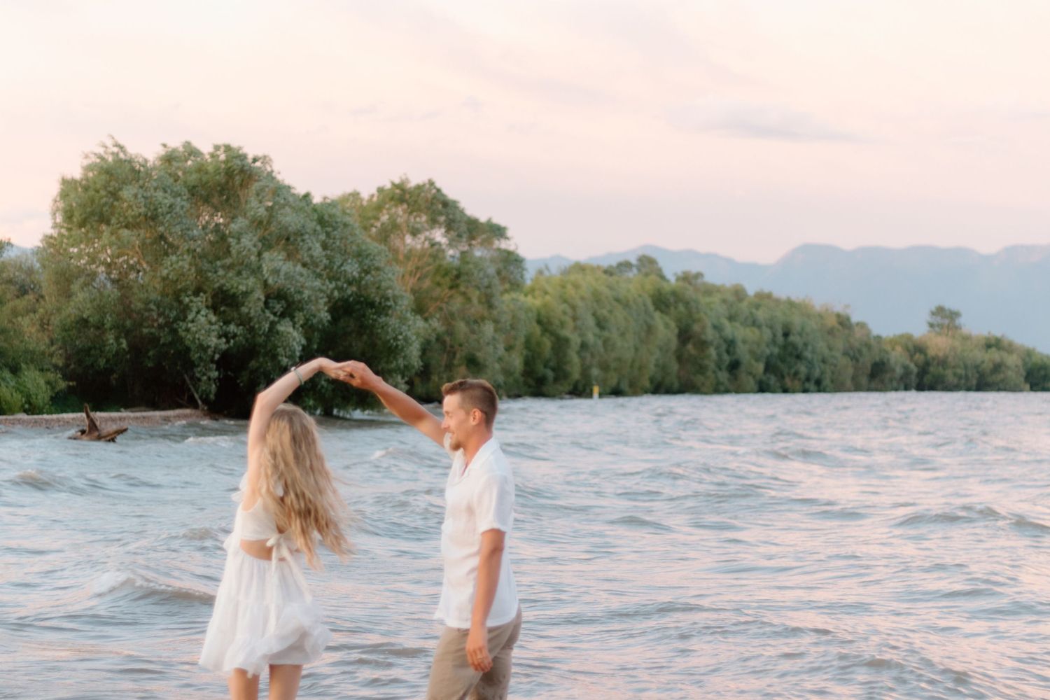 A couple stands together in shallow water at sunset with mountains and trees visible in the background.
