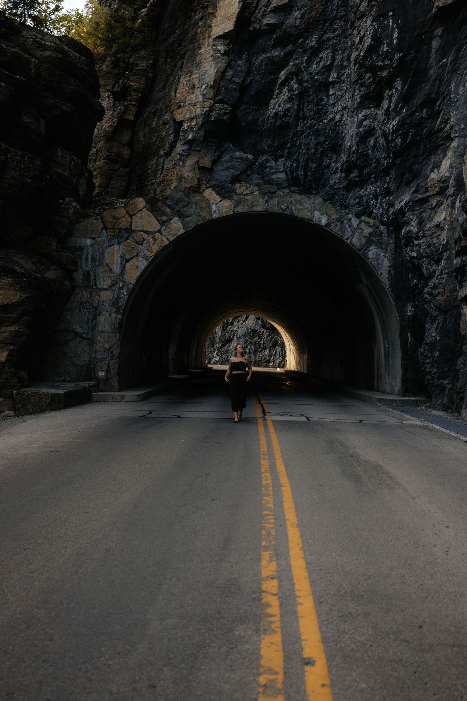 Dark tunnel carved through mountain rock with yellow road lines leading into distant light opening.