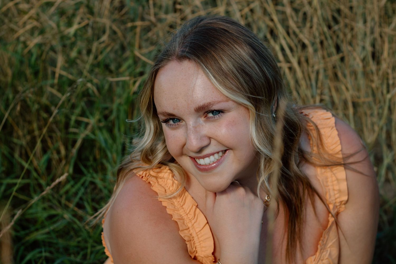 A woman in a peach-colored dress poses outdoors against a natural backdrop of tall grass and greenery.