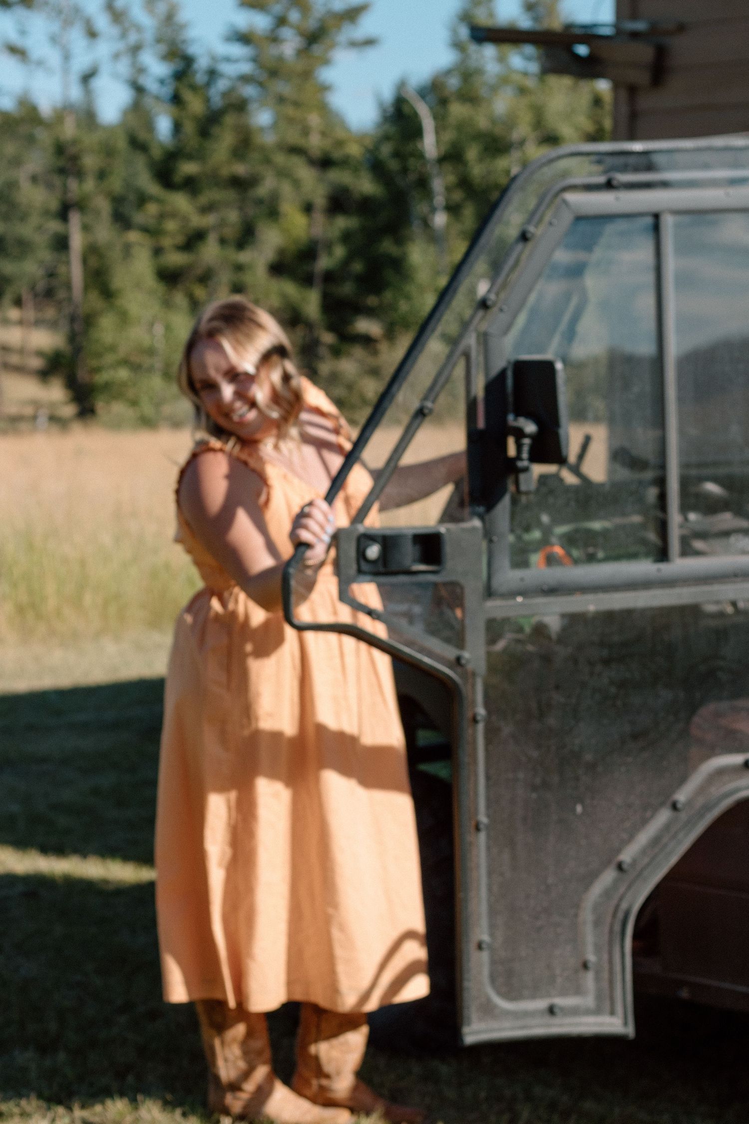 A person in a peach-colored dress stands next to a vintage Land Rover vehicle in a grassy outdoor setting.