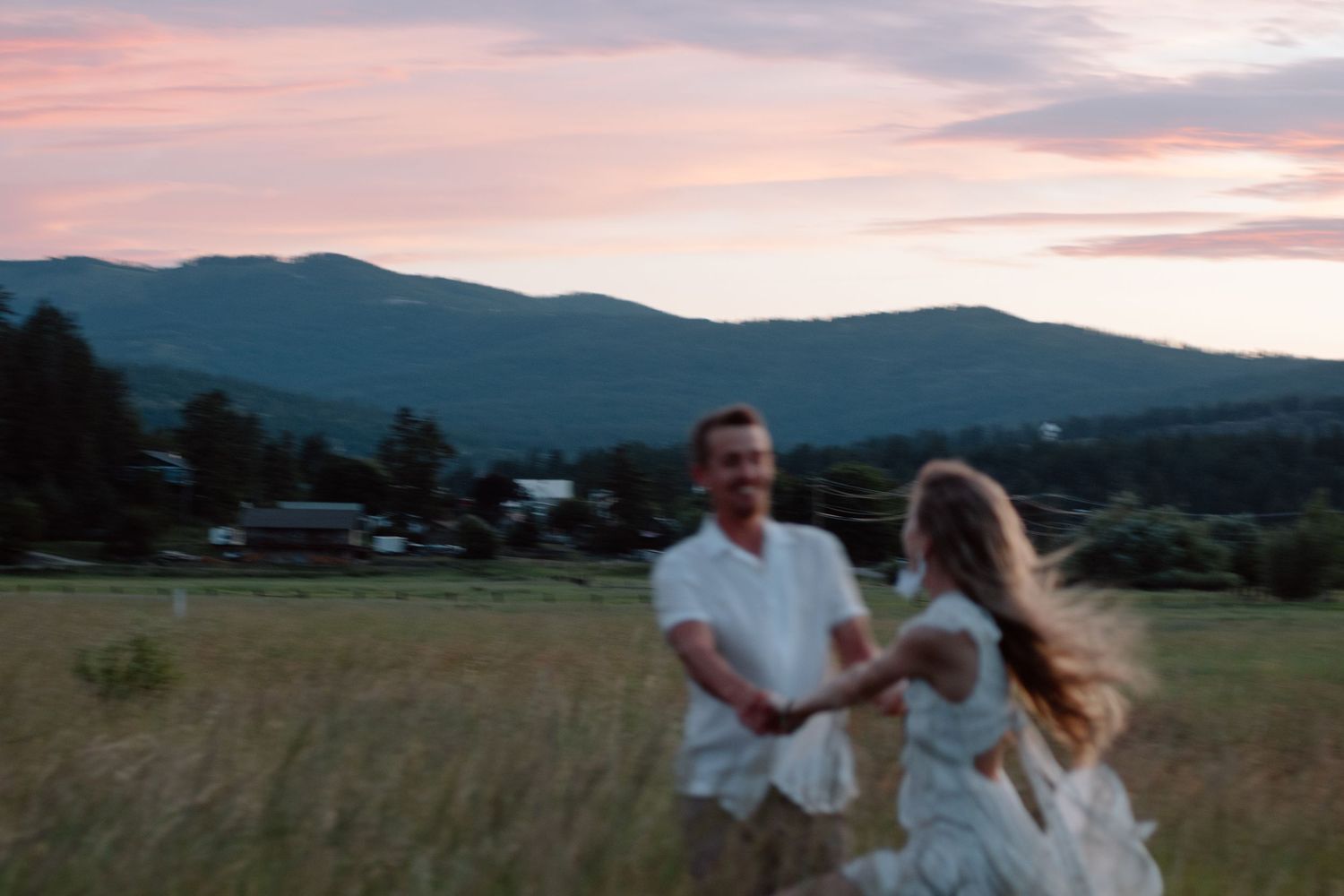 A couple dances together in a field with mountains visible in the background at sunset.