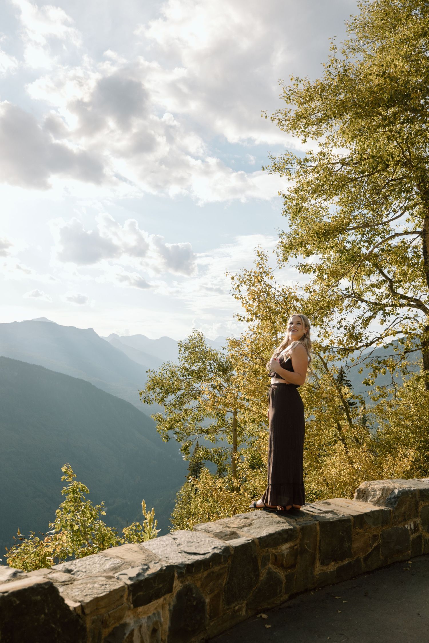 A person in a black dress stands on a stone overlook with mountains and autumn trees in the background.