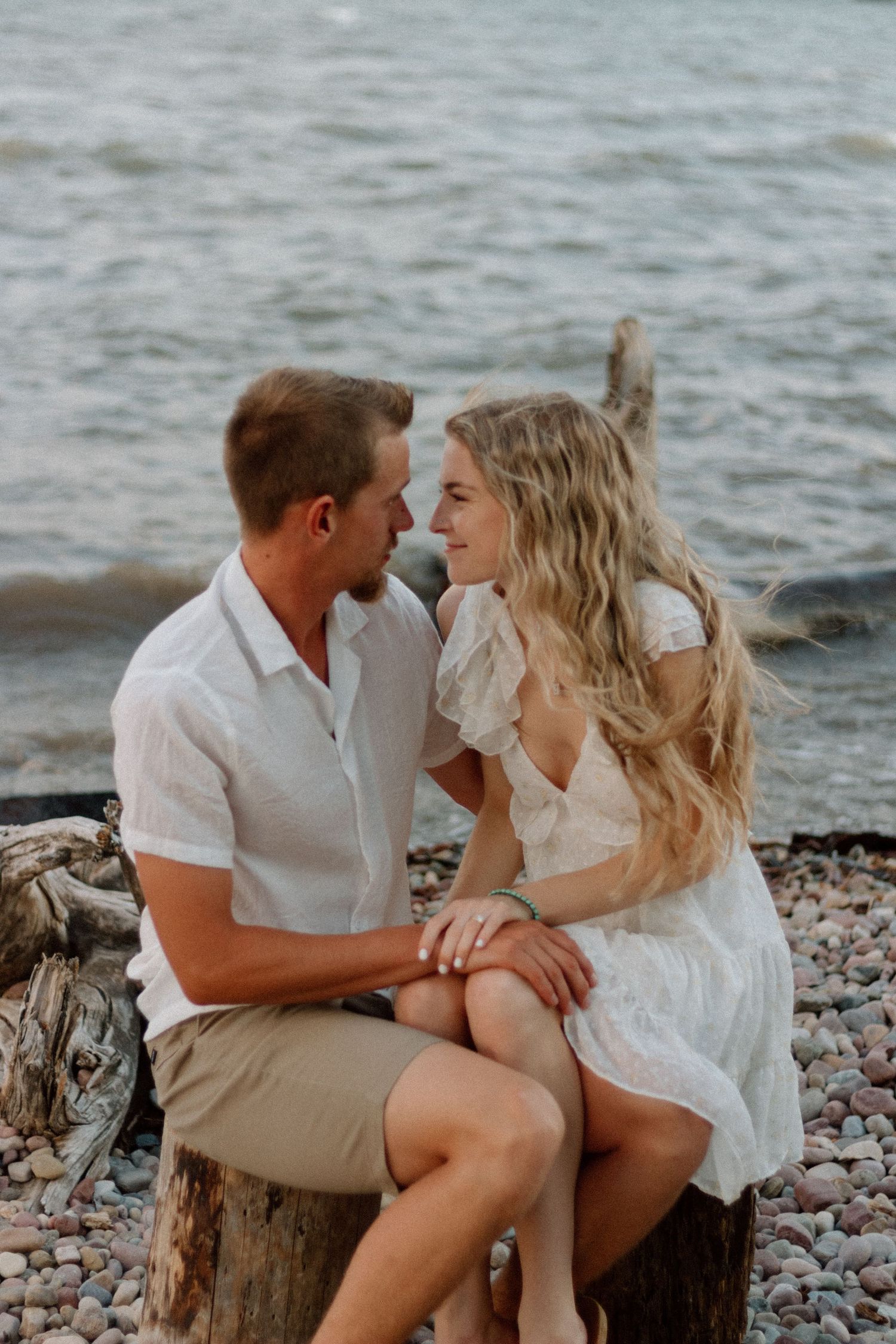 A romantic couple sits together on a rocky beach at sunset wearing white and beige outfits.