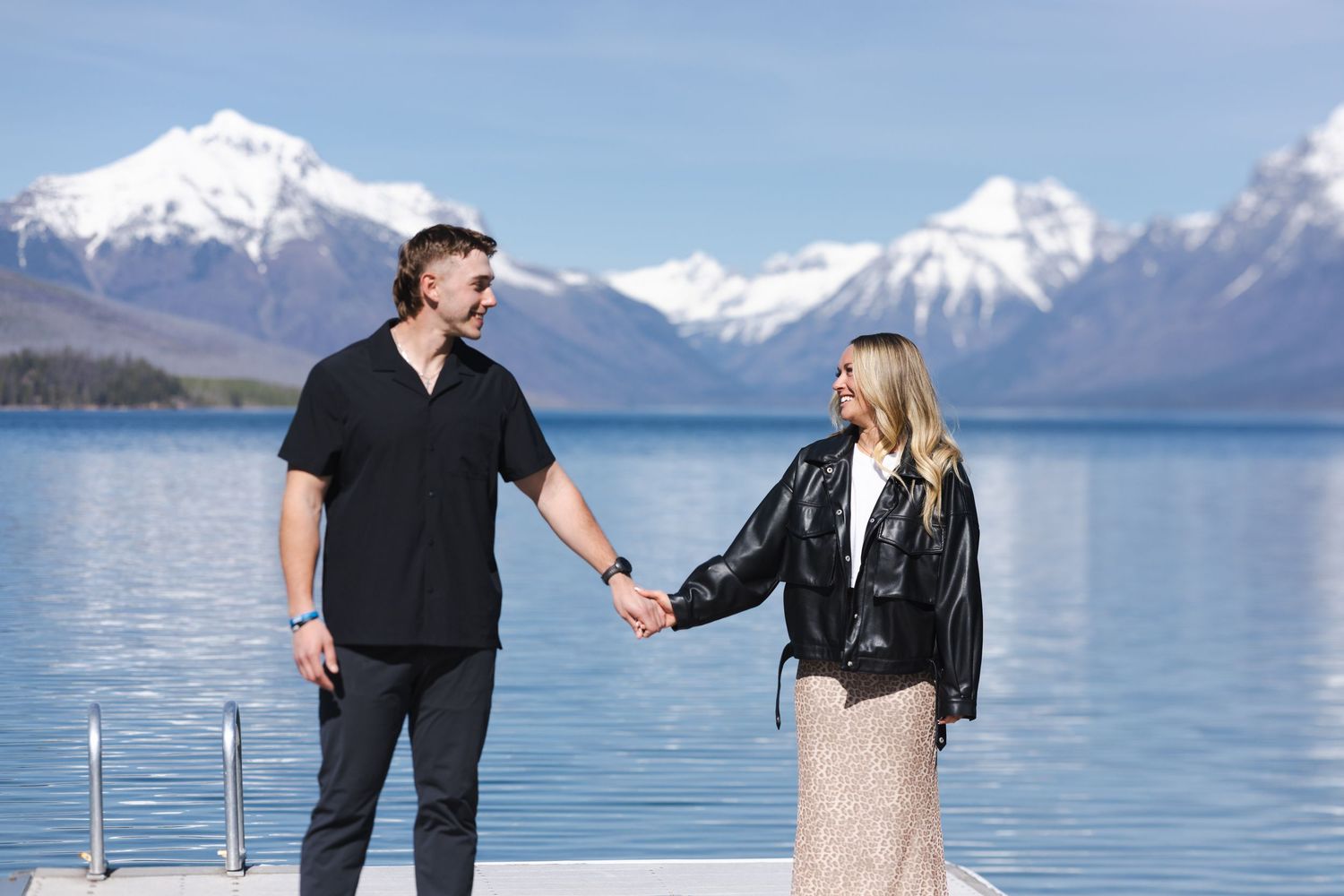 Couple holding hands on a dock with stunning snow-capped mountains and blue lake in background.