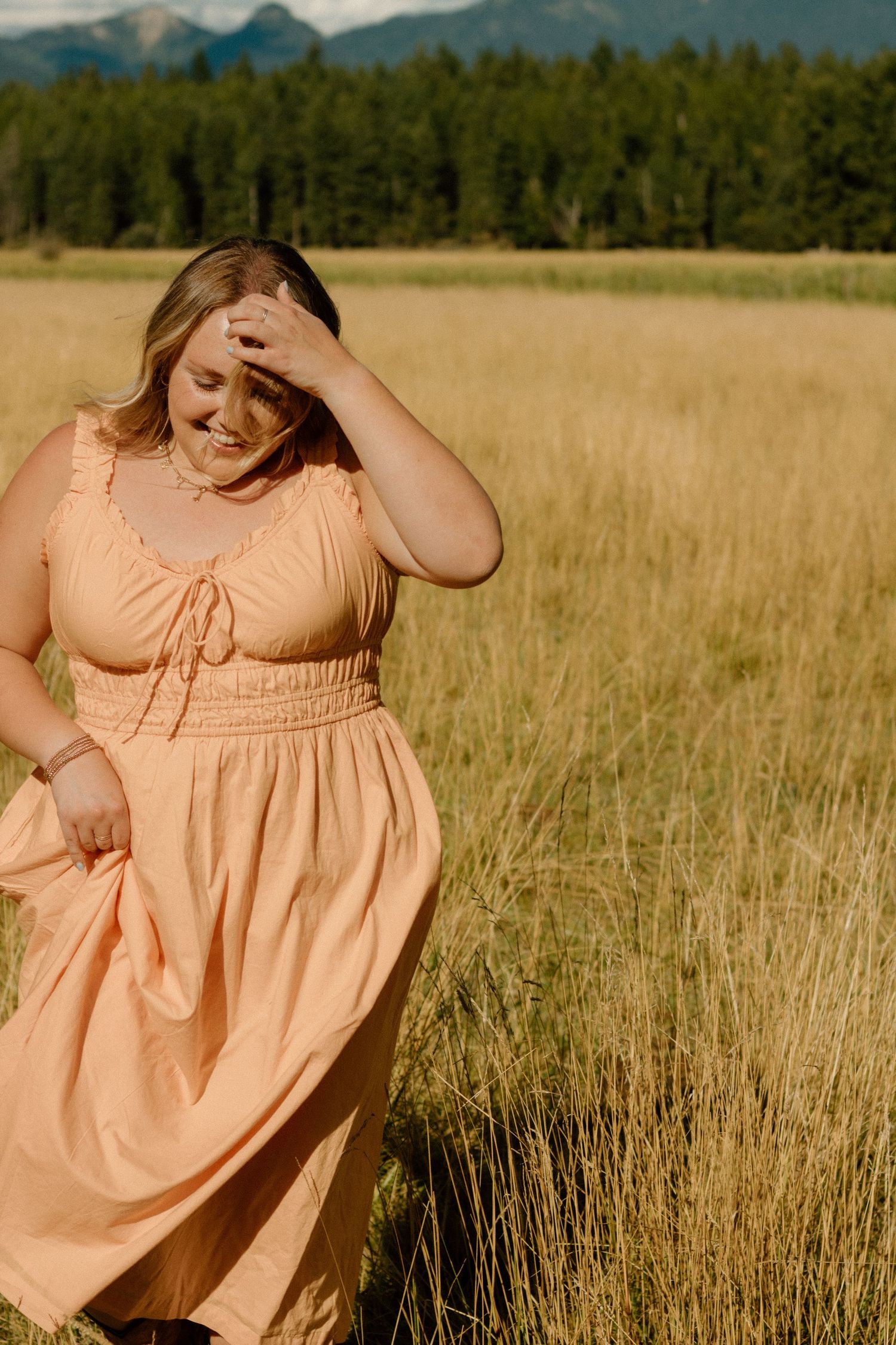 Peachy flowy dress movement captured in golden wheat field with mountain backdrop and forest in distance.