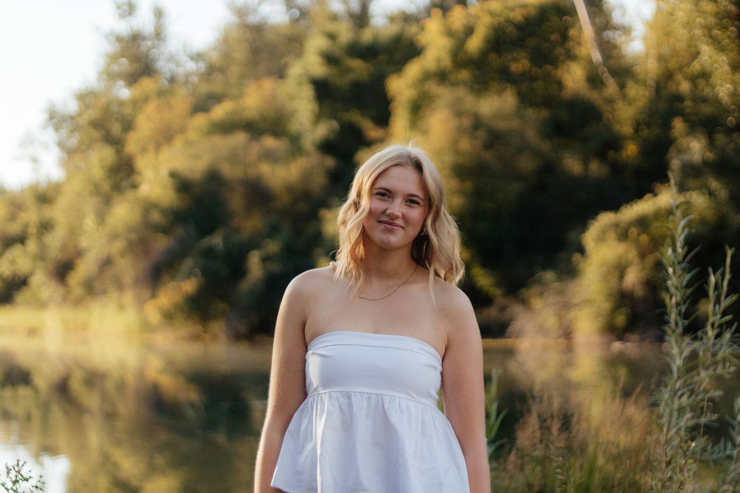 A person in a white dress poses outdoors during golden hour in a natural setting with warm sunlight filtering through trees.