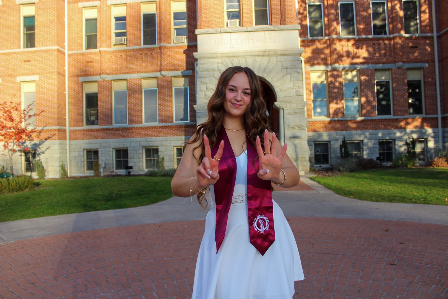Student walking across sunny collegiate campus quad near red brick buildings wearing pink dress in autumn.