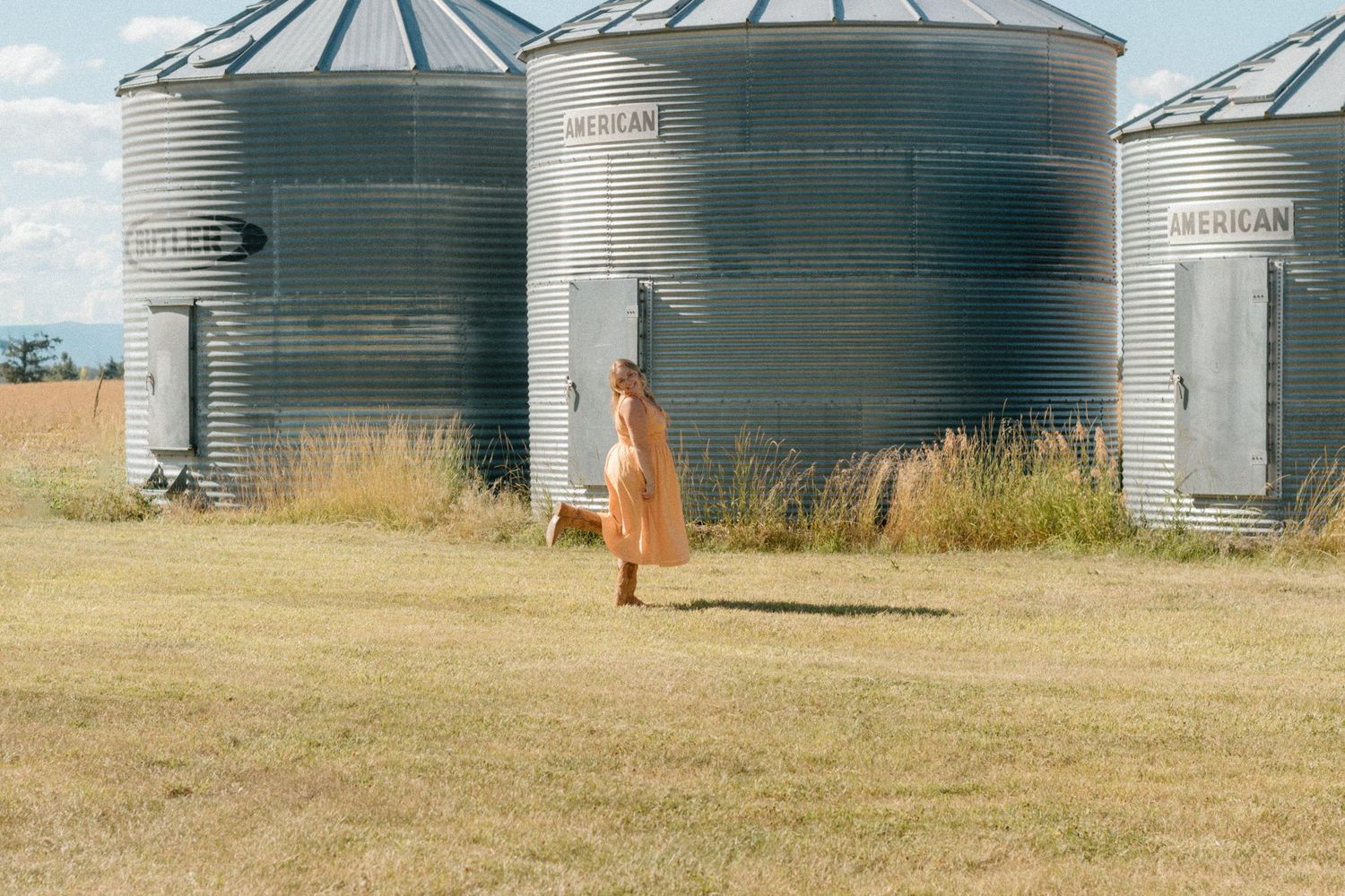Person standing near large metal grain storage bins at a rural farm facility under cloudy skies.