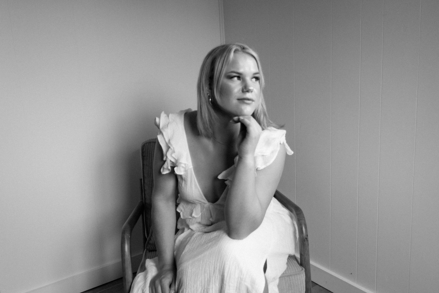 A black and white photo series shows a woman in a white dress sitting and posing against a plain wall.