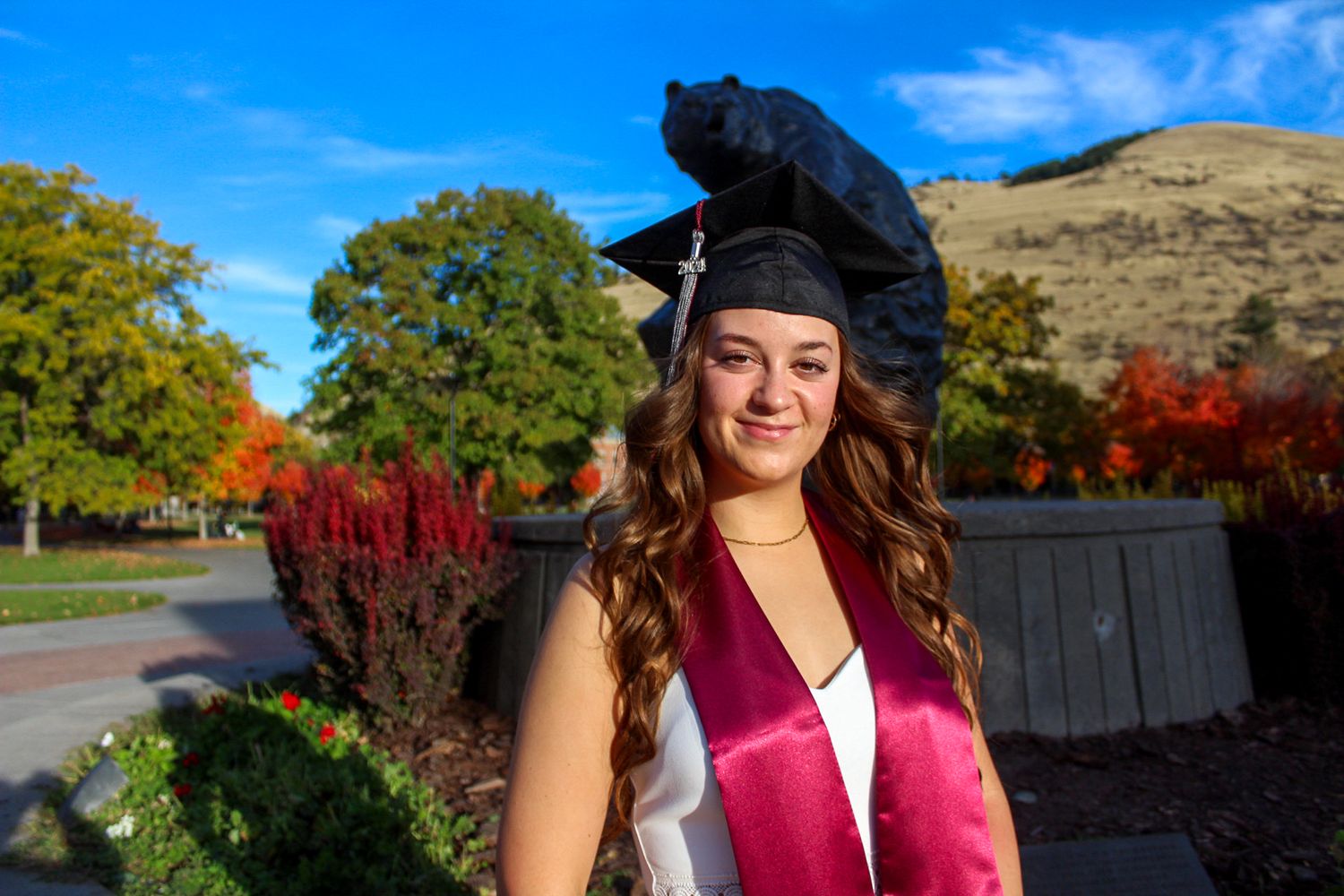 Five graduating students in academic regalia pose outdoors against autumn foliage and campus buildings.