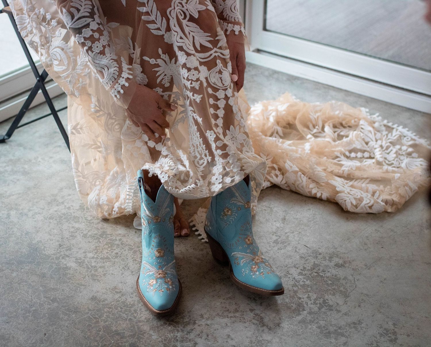 Light blue cowboy boots paired with a flowing lace wedding dress on concrete floor.