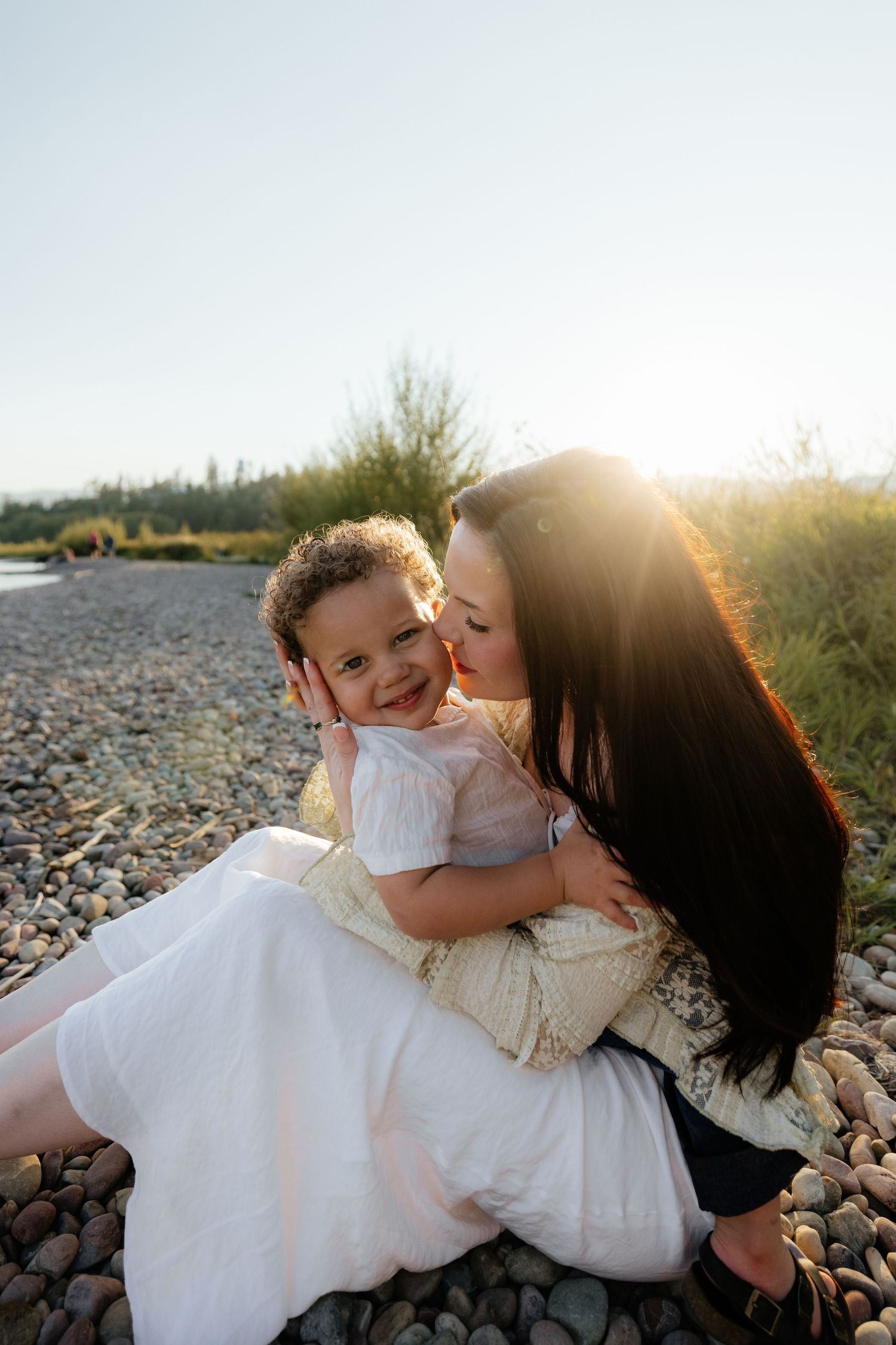 Mother and daughter share tender moments on a rocky beach at sunset with warm golden light glowing behind them.