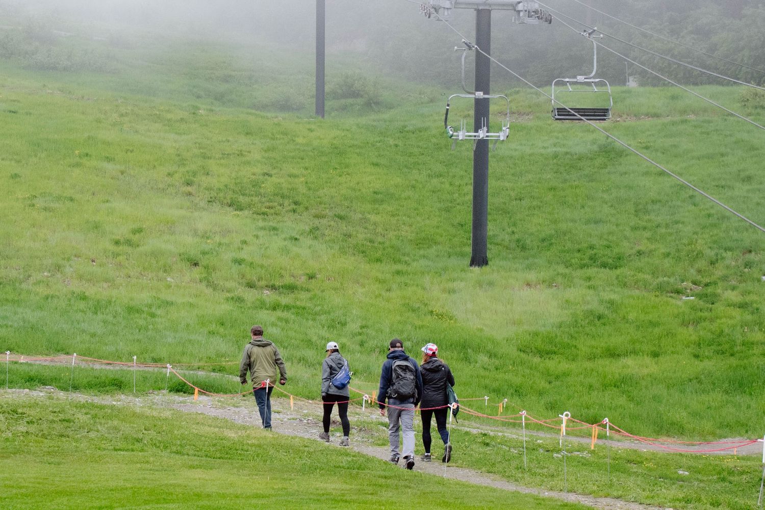 Hiking trail on green mountain slope with chairlift towers and distant hikers visible.