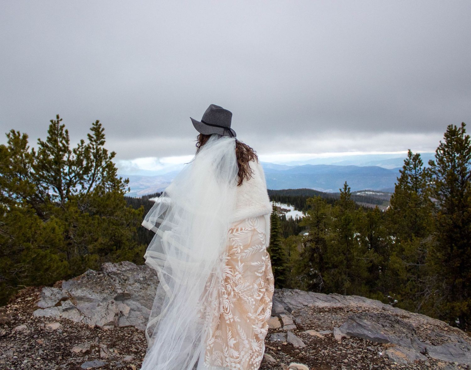 A bride in a flowing white dress stands on a rocky mountain overlook with scenic mountain vistas in the background.