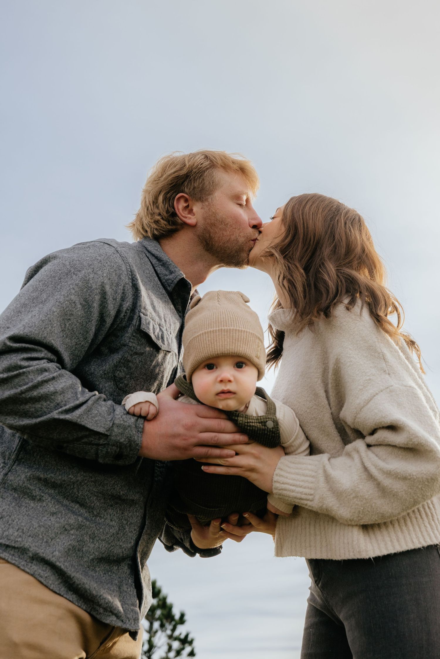 A couple shares tender kisses while holding their baby in a series of intimate outdoor family photos.