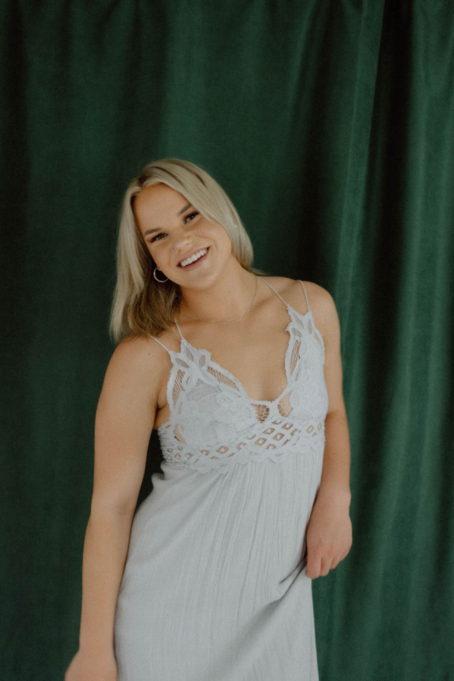 Woman in white lace-trimmed camisole poses against green curtain backdrop.