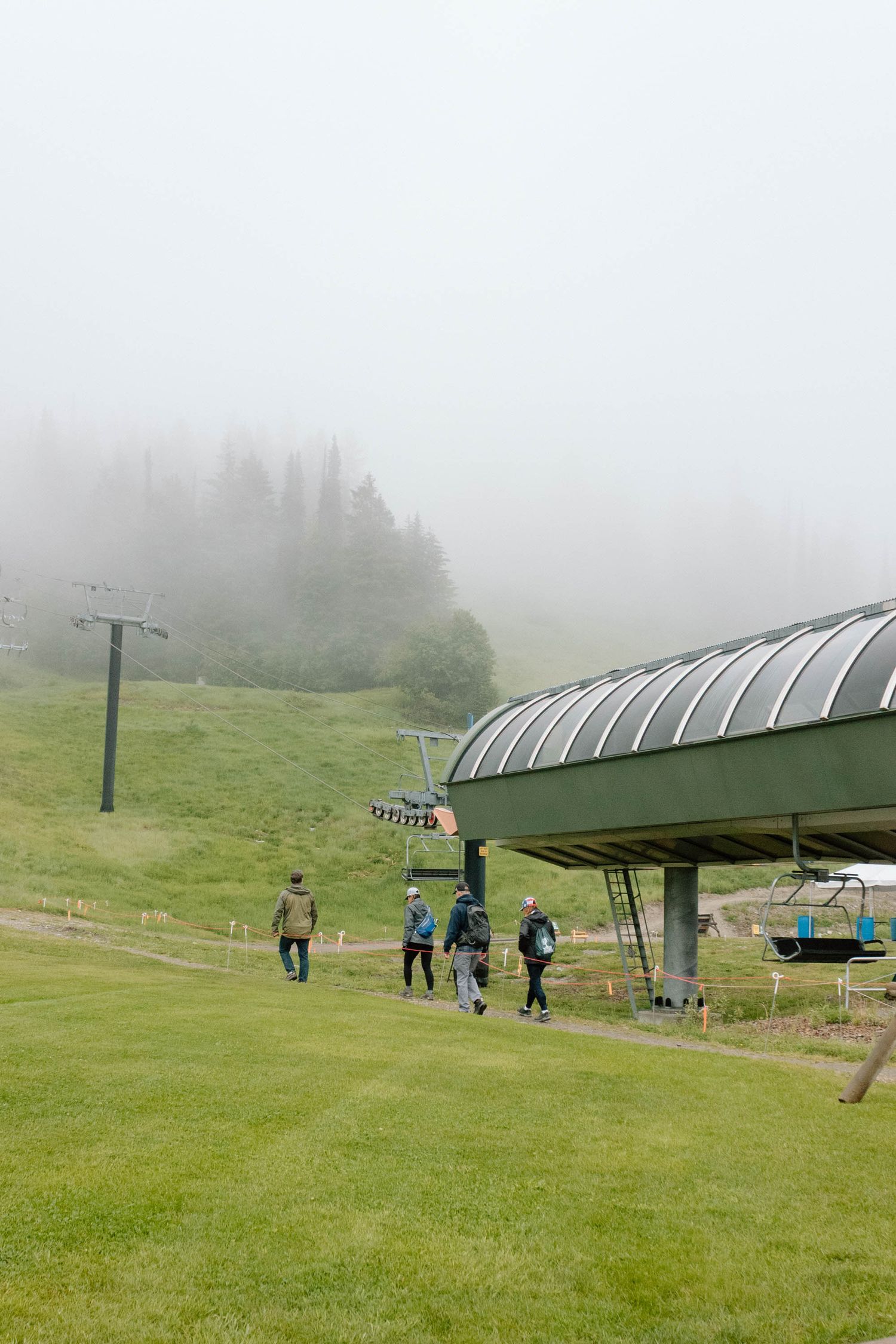 A misty ski lift on a green mountain slope with distant figures walking beneath.