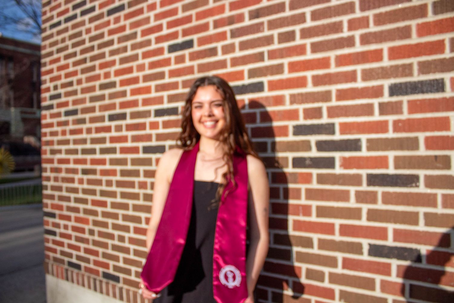 Person in magenta blazer stands against red brick wall on sunny day.
