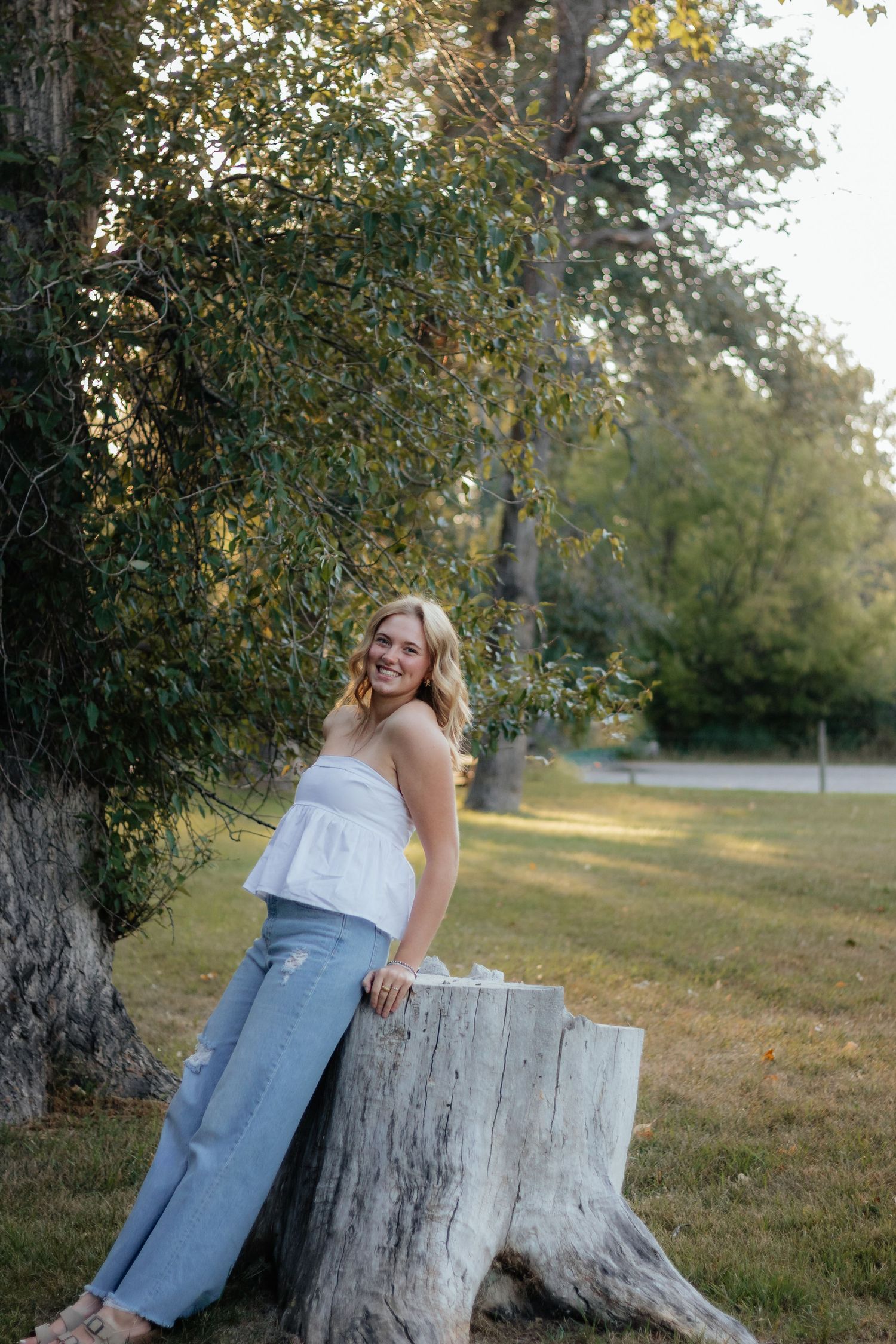 A person in a white top and jeans poses casually against a weathered tree stump in a sunlit outdoor setting.