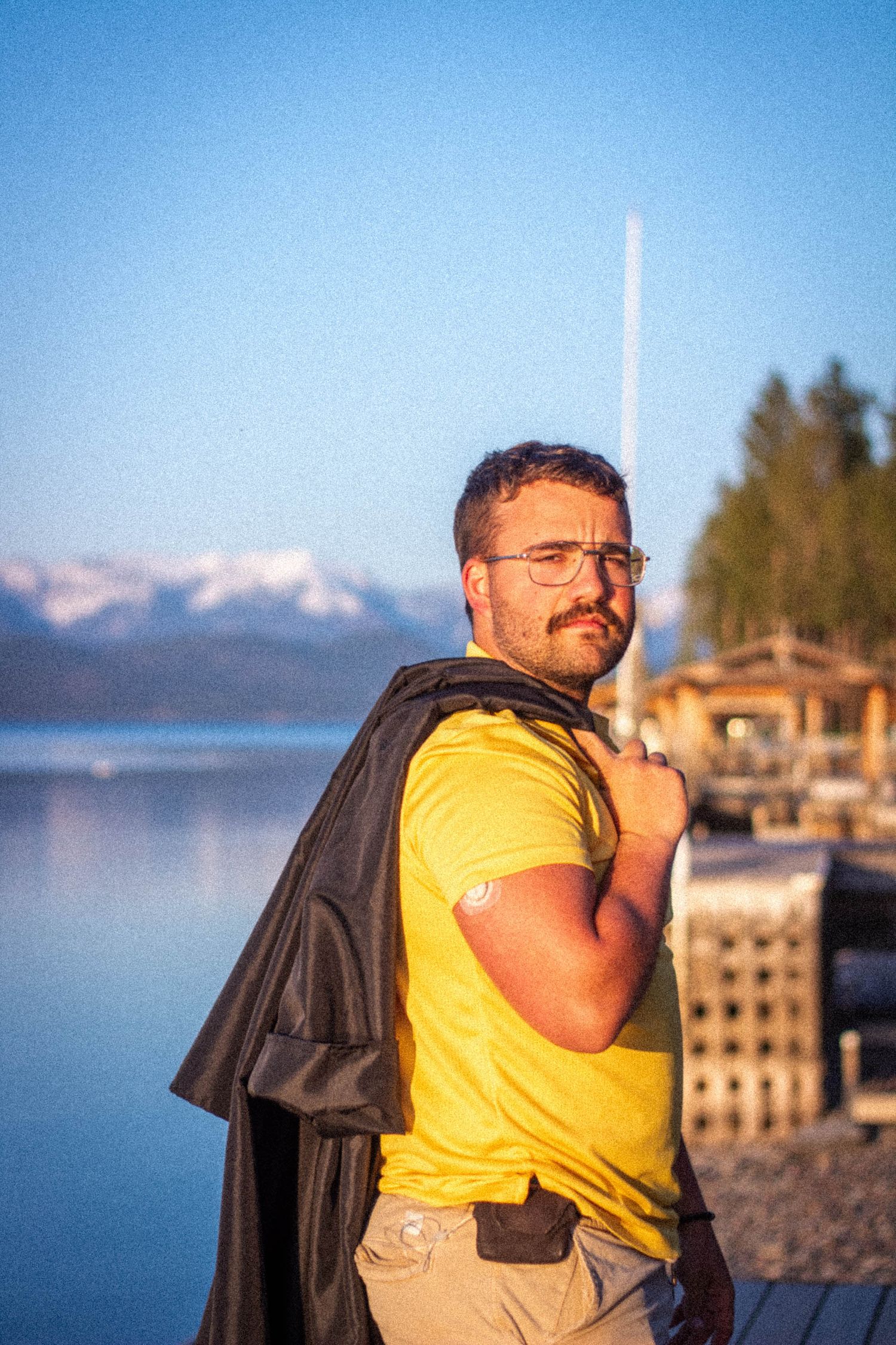 Person in yellow t-shirt stands at a lakeside dock with mountains visible in the background during golden hour.