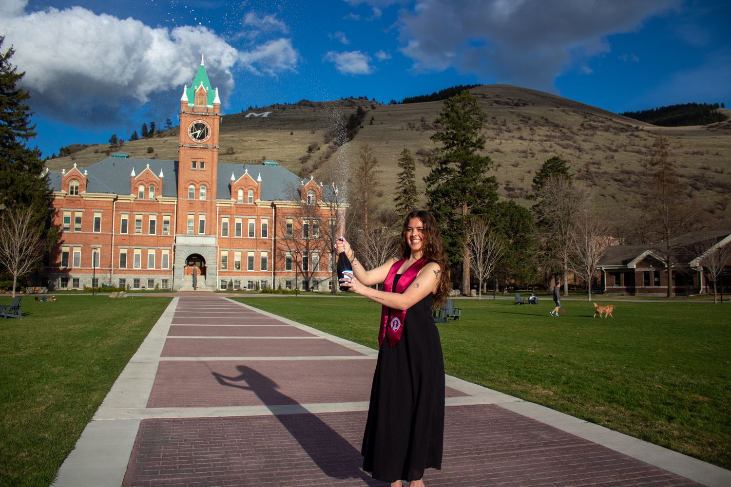 Graduate in black dress celebrates commencement in front of University Main Hall with mountains in background.