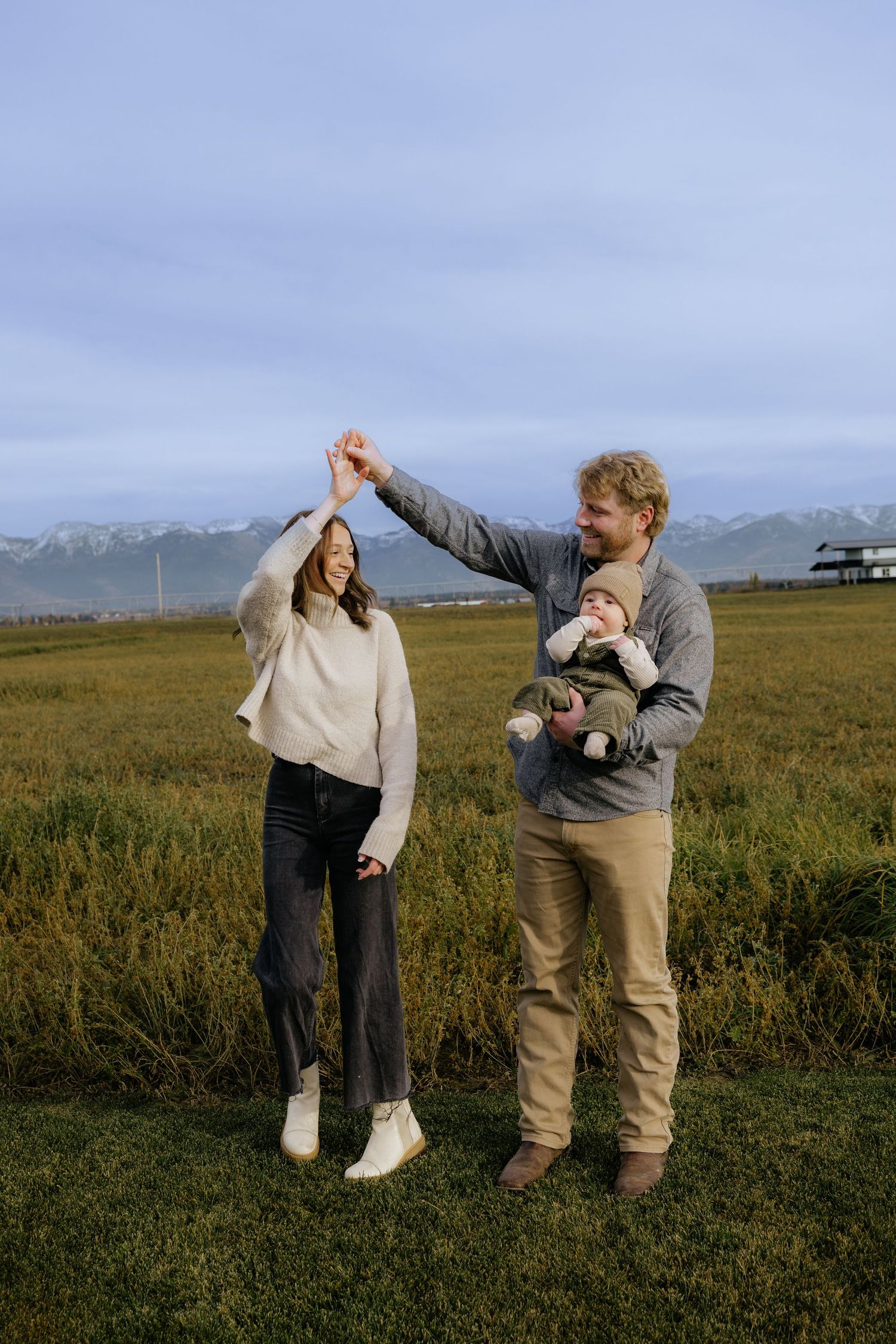 A couple dances playfully in a grassy field with mountains visible in the background on a cloudy day.