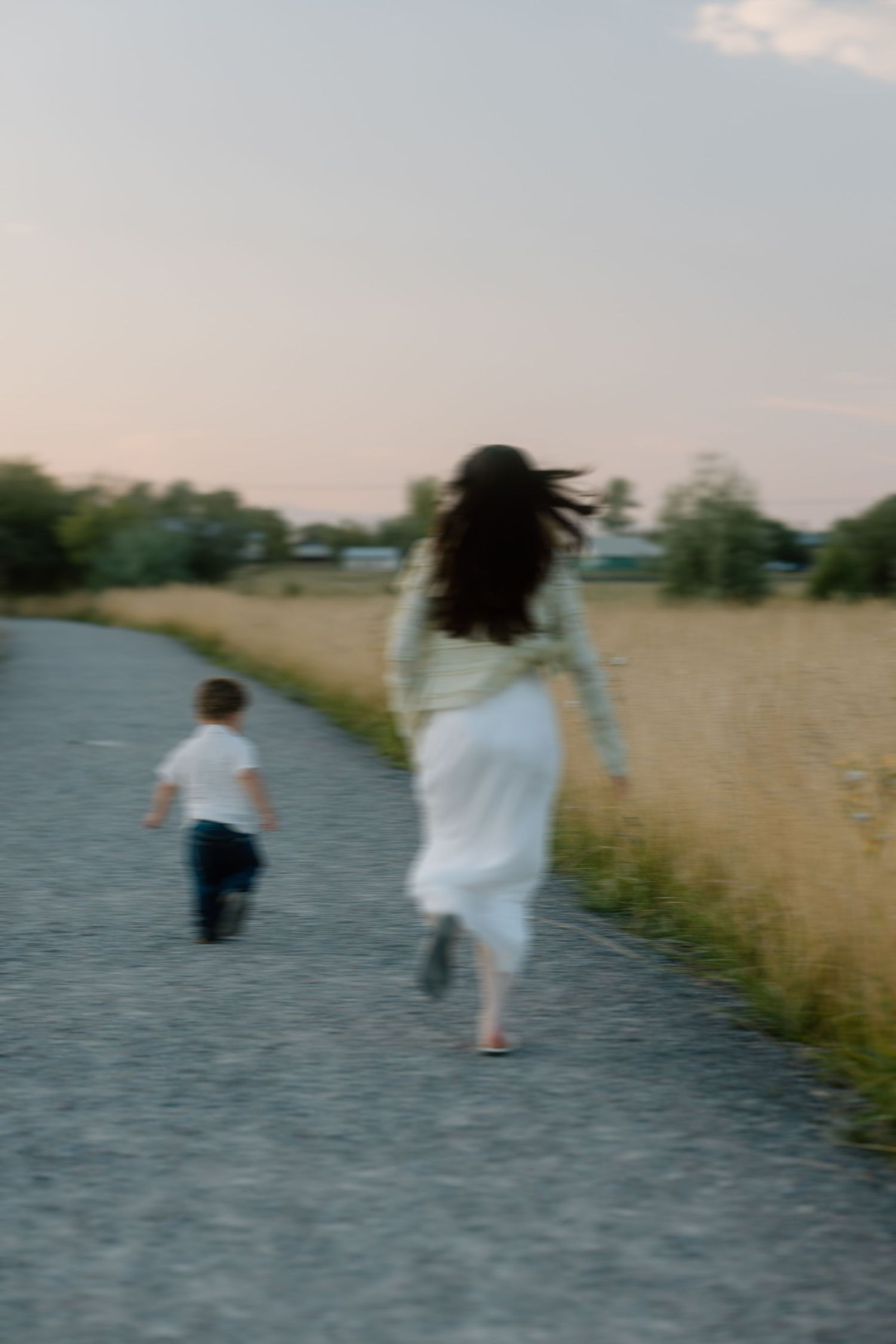 Two people walk along a gravel path beside tall grass fields at dusk.