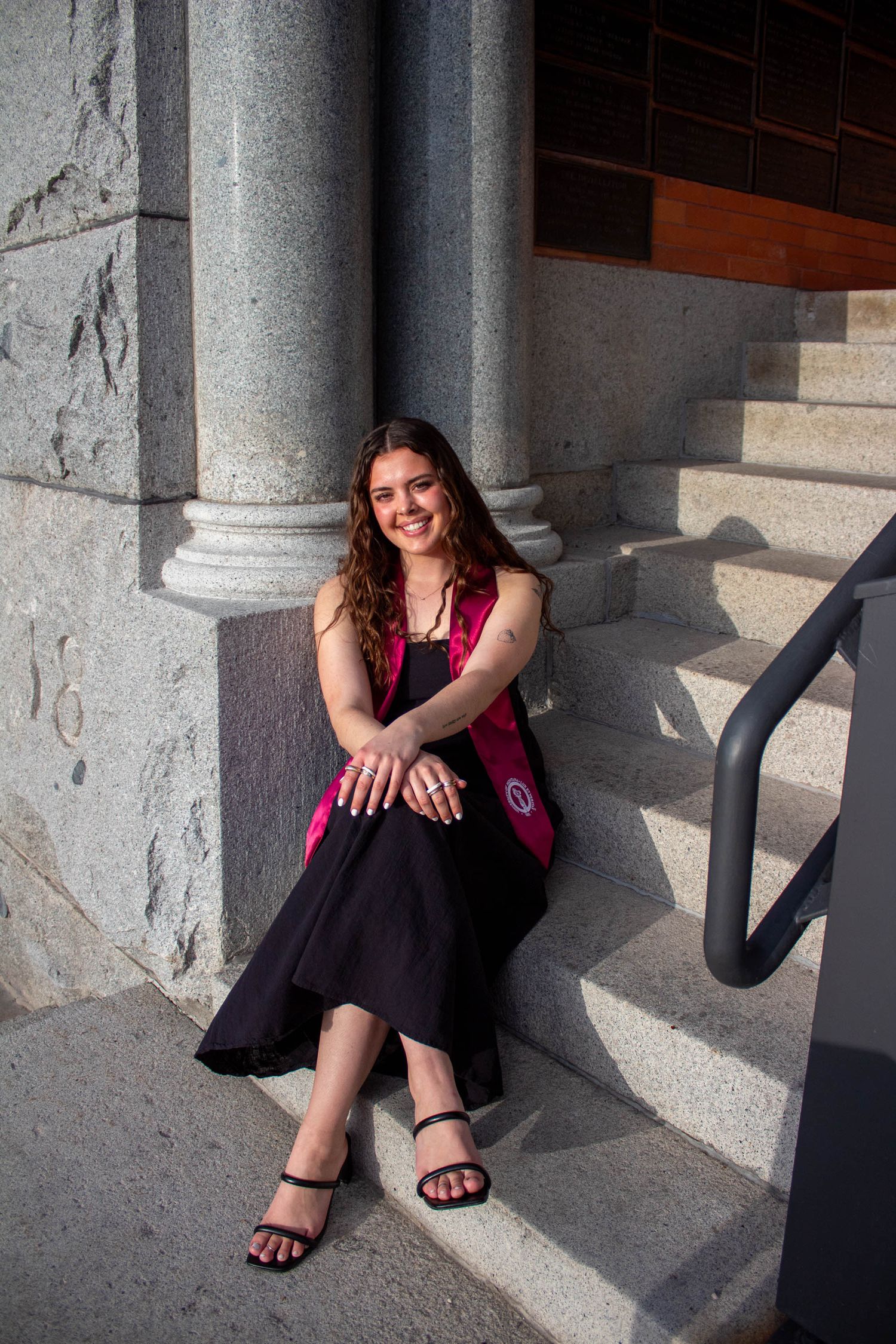 A person in a red tank top and black skirt sits casually against stone columns while casting shadows in dramatic lighting.