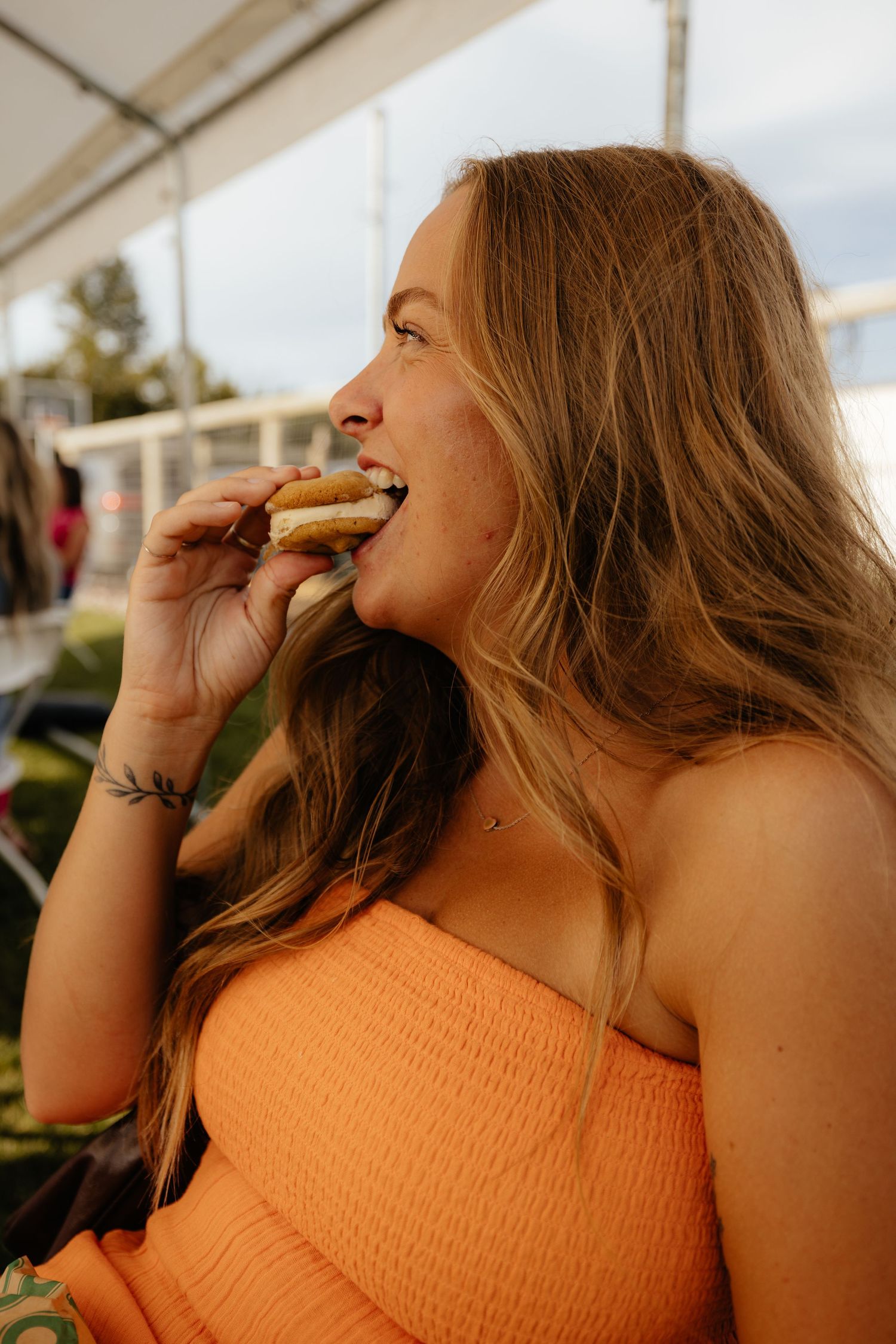 Person in orange top enjoying food at an outdoor event.