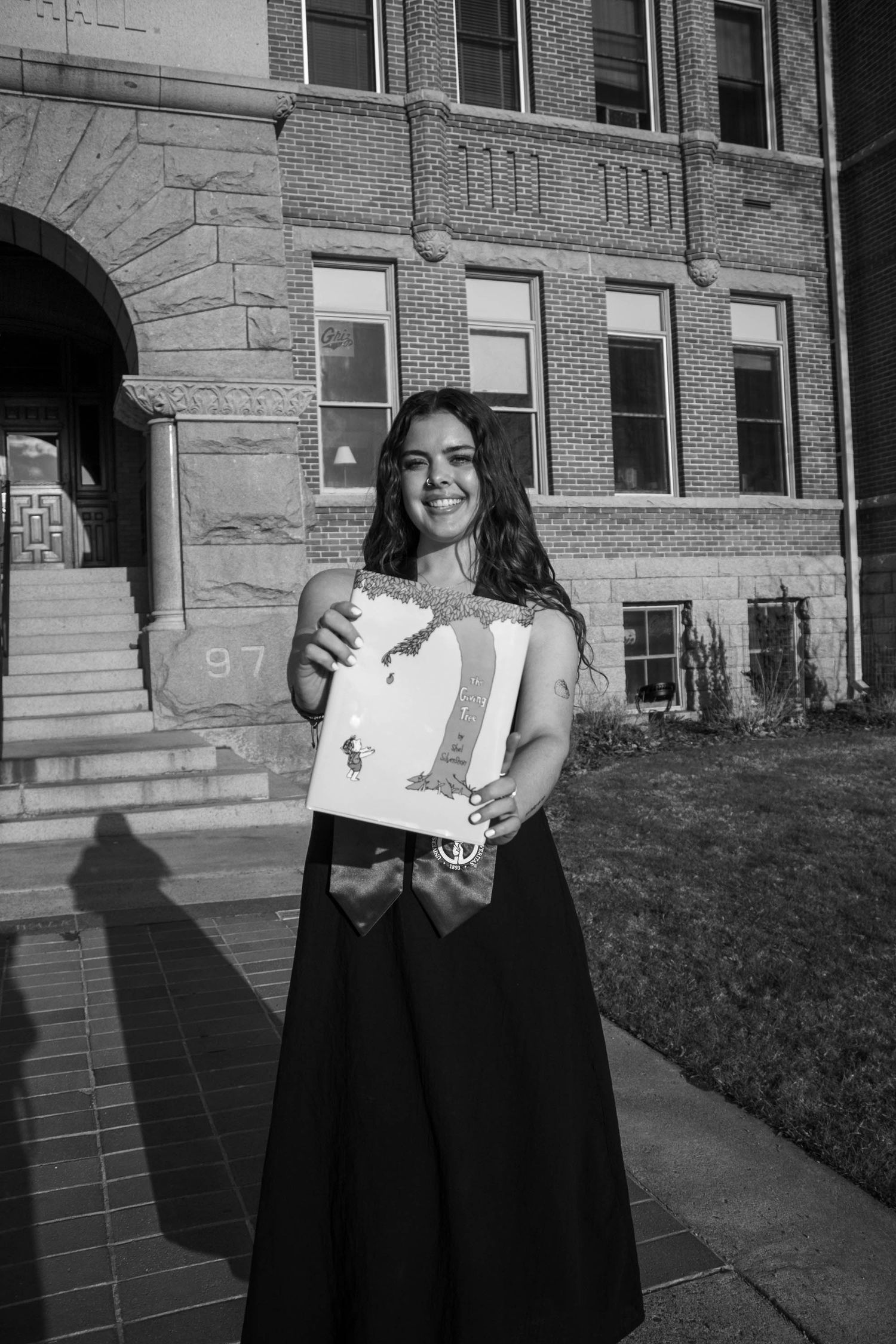A graduate stands outside a brick building wearing a long black skirt while holding a diploma and a beverage.