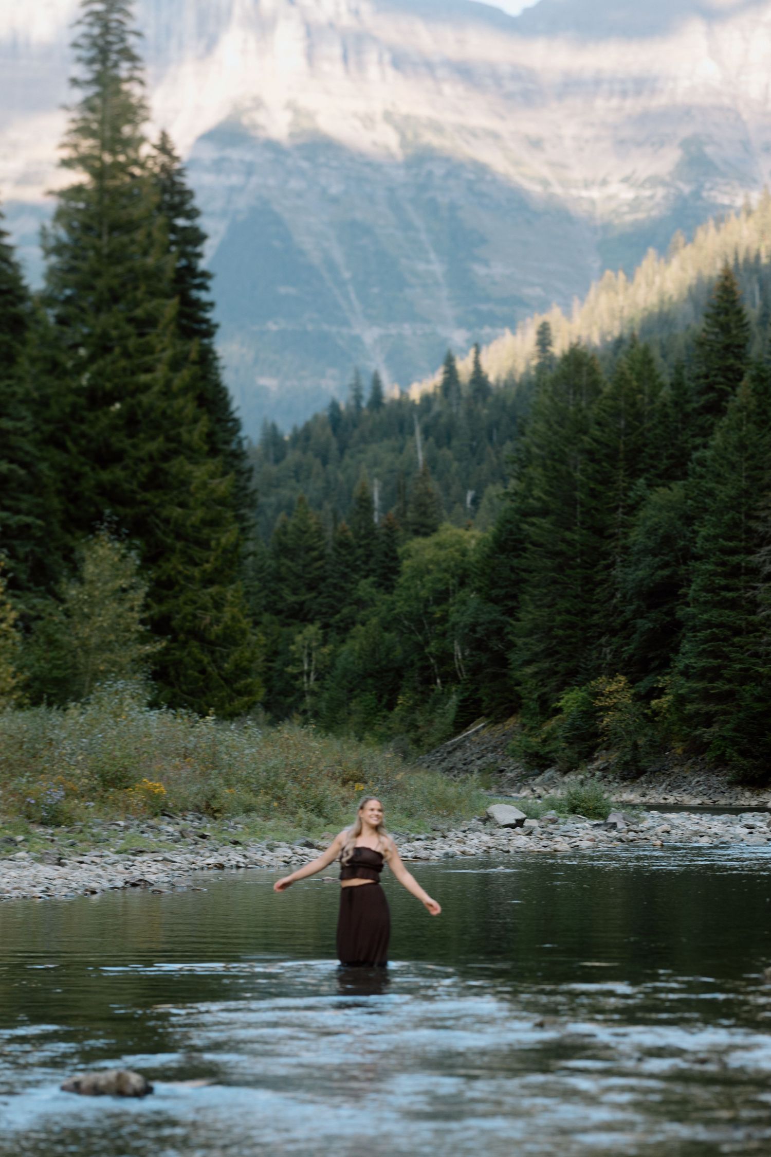 A figure wades in a serene mountain lake surrounded by evergreen forests and snow-capped peaks.