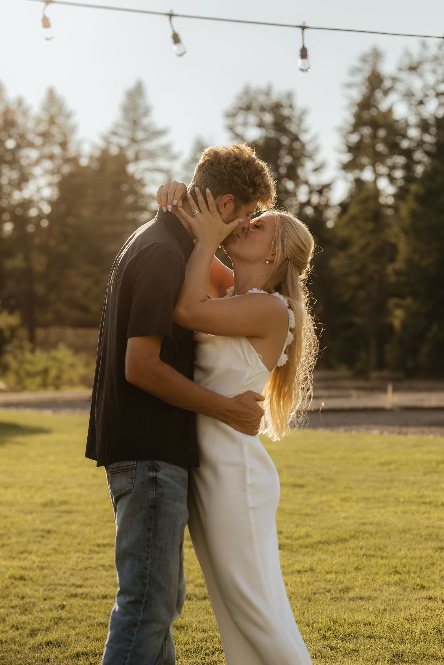 A couple shares an intimate embrace in a sun-drenched field during golden hour with evergreen trees in the background.